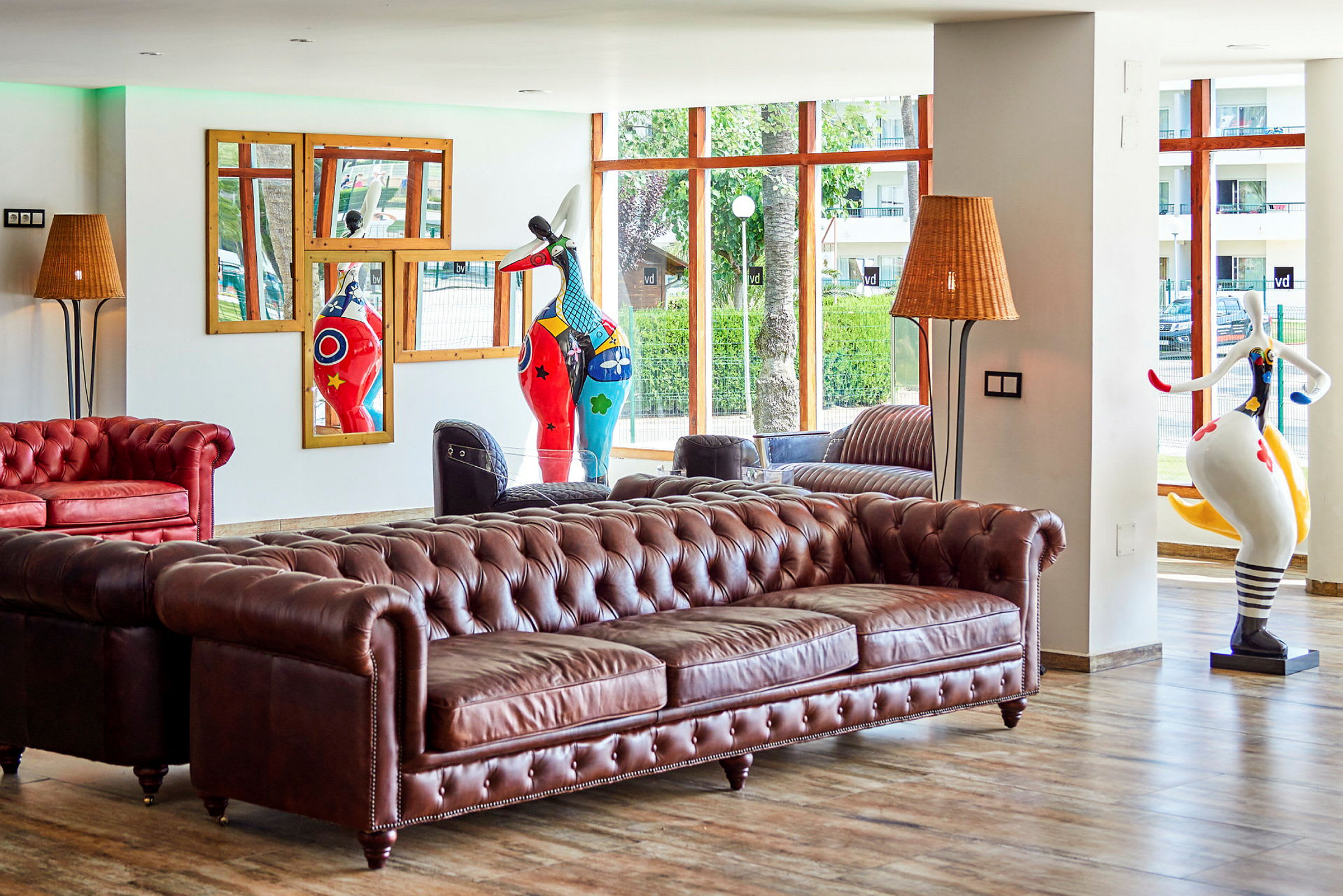 En hotellobby med en brun chesterfield-sofa, dekorative skulpturer og store vinduer med udsigt. (AI-genereret tekst)