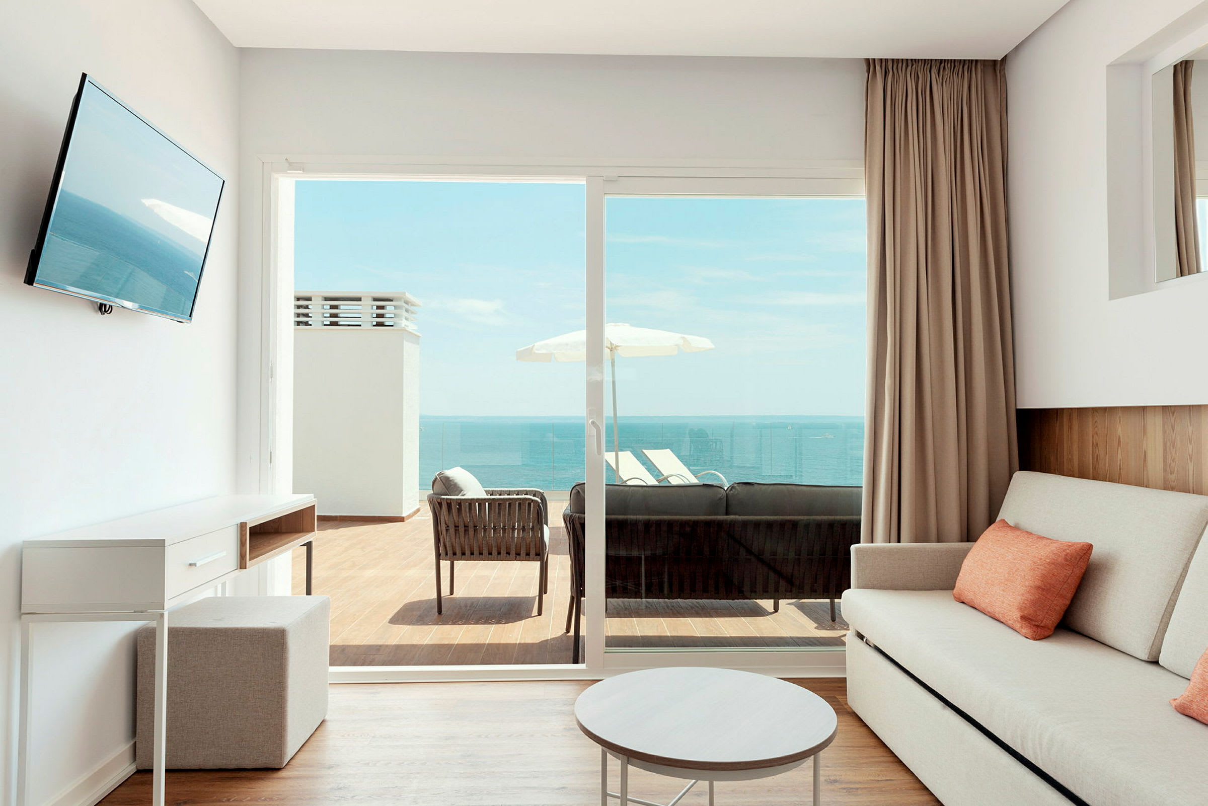 Et lyst rum med en sofa og bord, som har en udendørs balkon med siddepladser og udsigt over havet. (AI-genereret tekst)