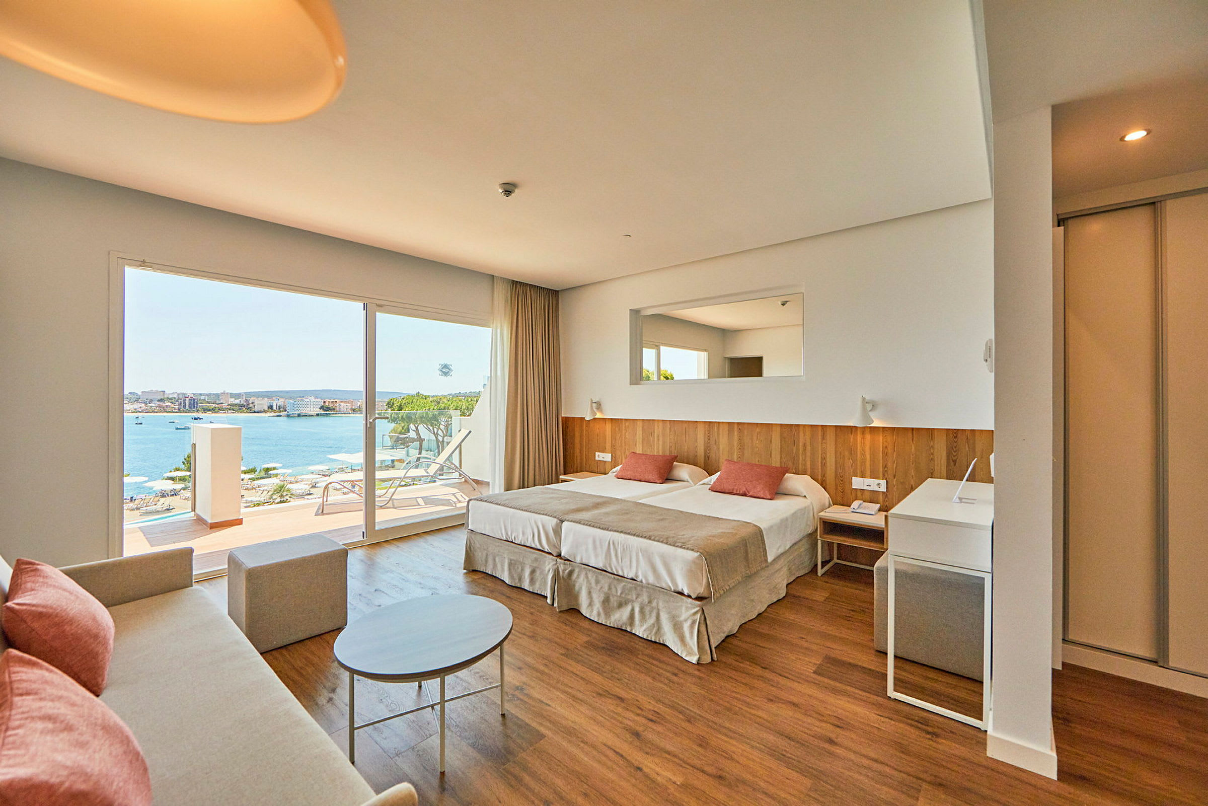 Et hotelværelse med dobbeltseng, sofa og store vinduer, der vender ud mod en terrasse med udsigt over havet. (AI-genereret tekst)