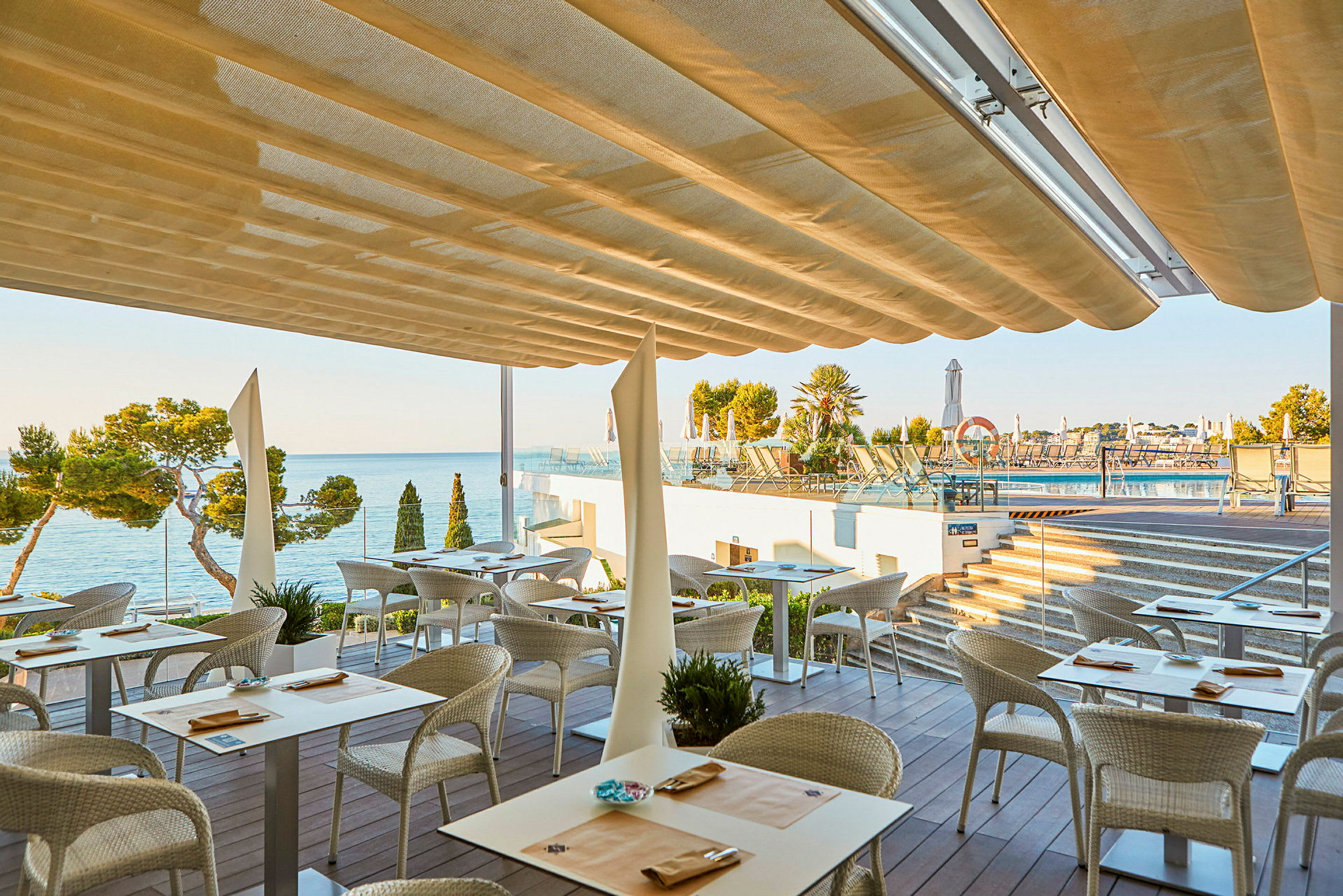 En restaurant med udendørs siddepladser under en markise med udsigt over havet og en pool. (AI-genereret tekst)