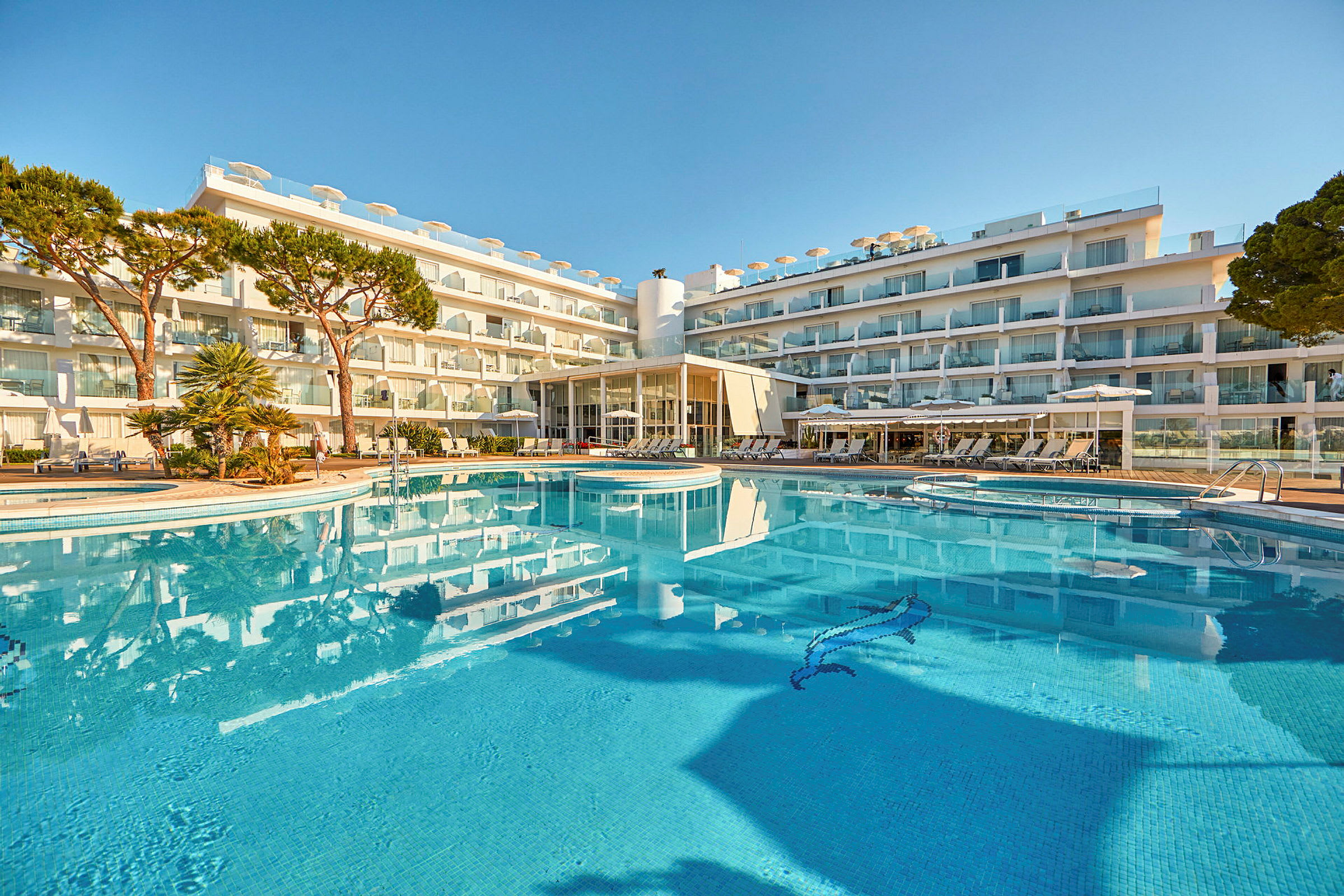 Et fler-etagers hotel med en udendørs pool omgivet af liggestole og grønne områder. (AI-genereret tekst)