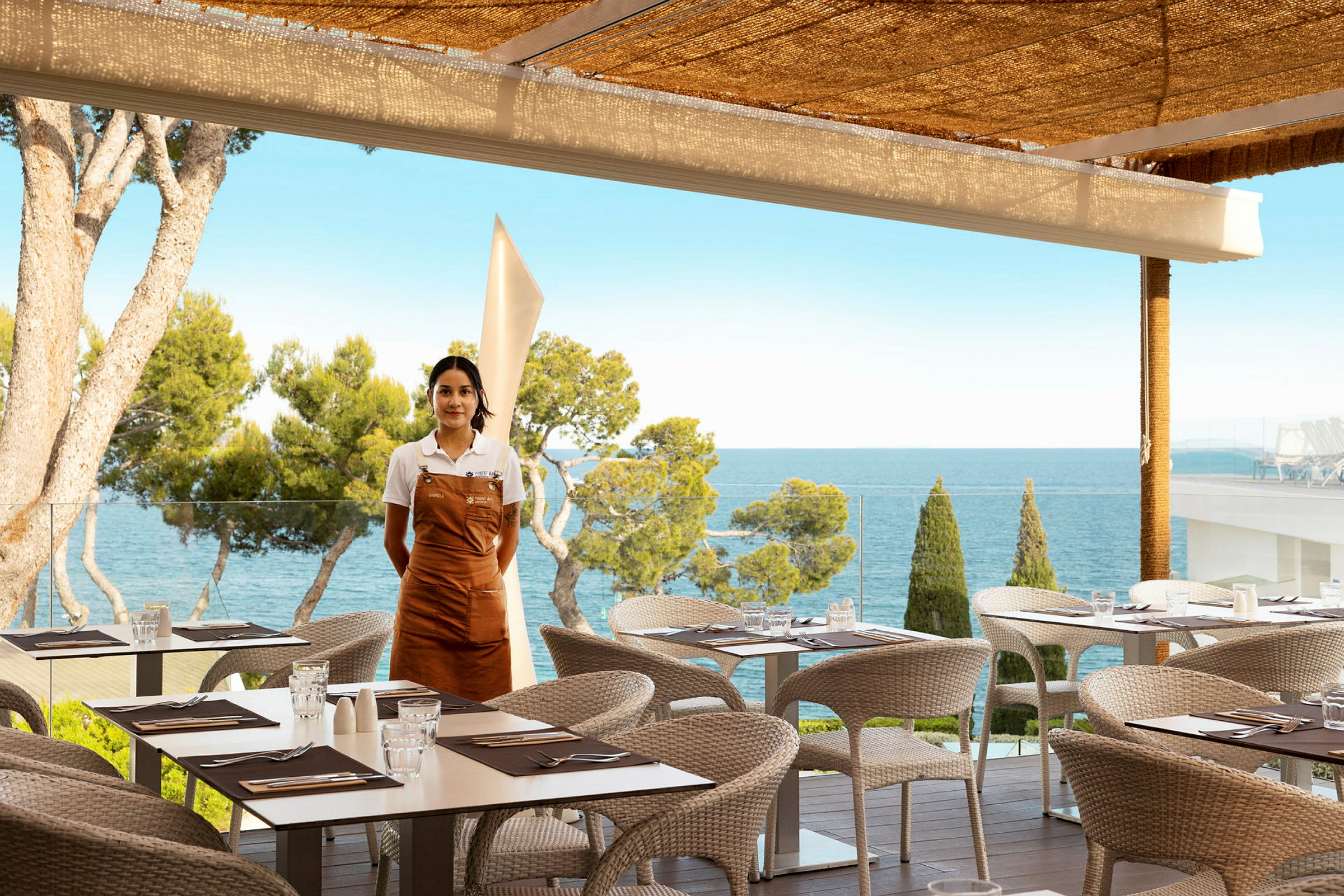 Restaurant med udendørs servering og havudsigt, en tjener står ved siden af. (AI-genereret tekst)