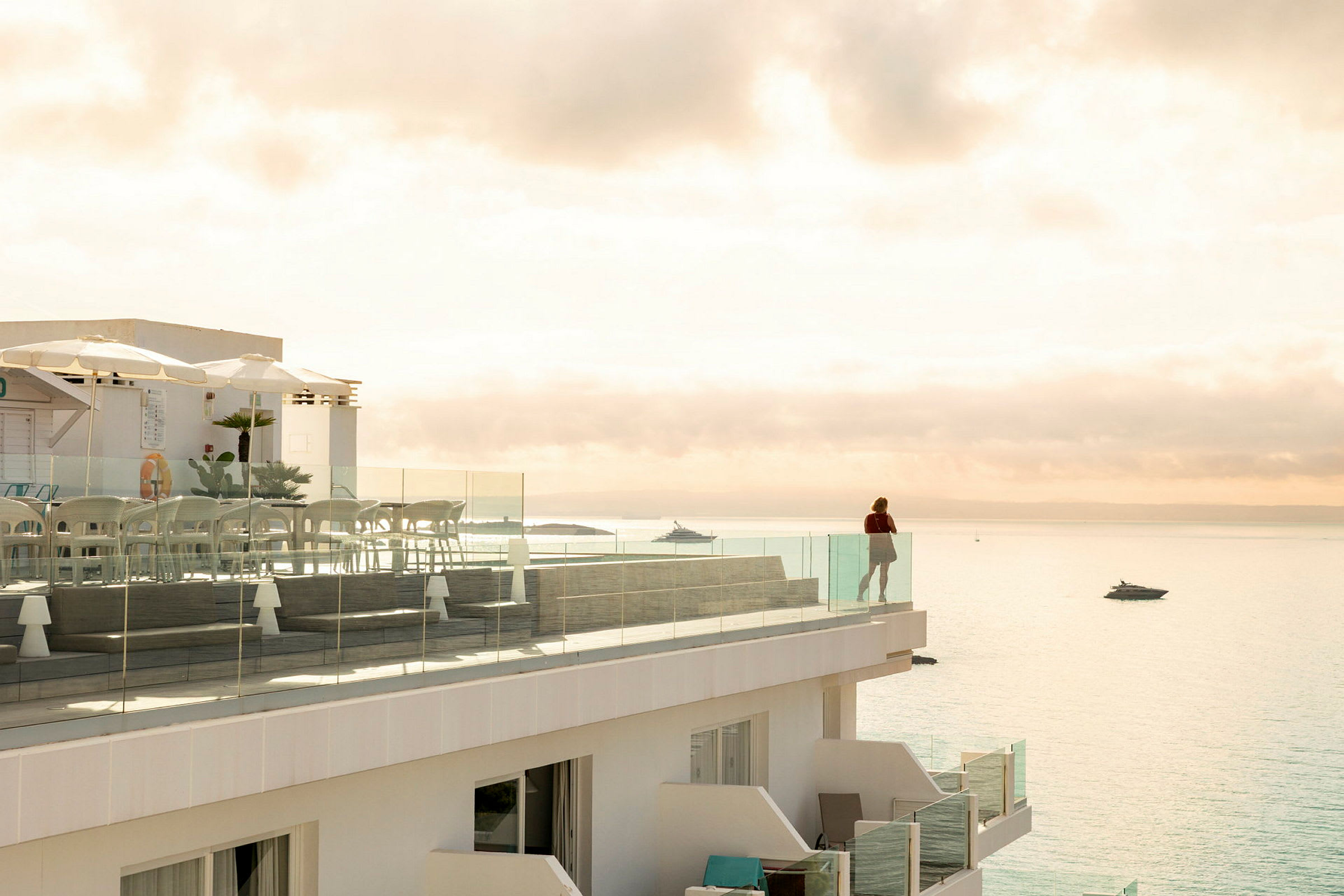En terrasse med glasrækværk, der har udsigt over havet. På terrassen står der stole og borde, og en person står ved rækværket og kigger ud over vandet. (AI-genereret tekst)