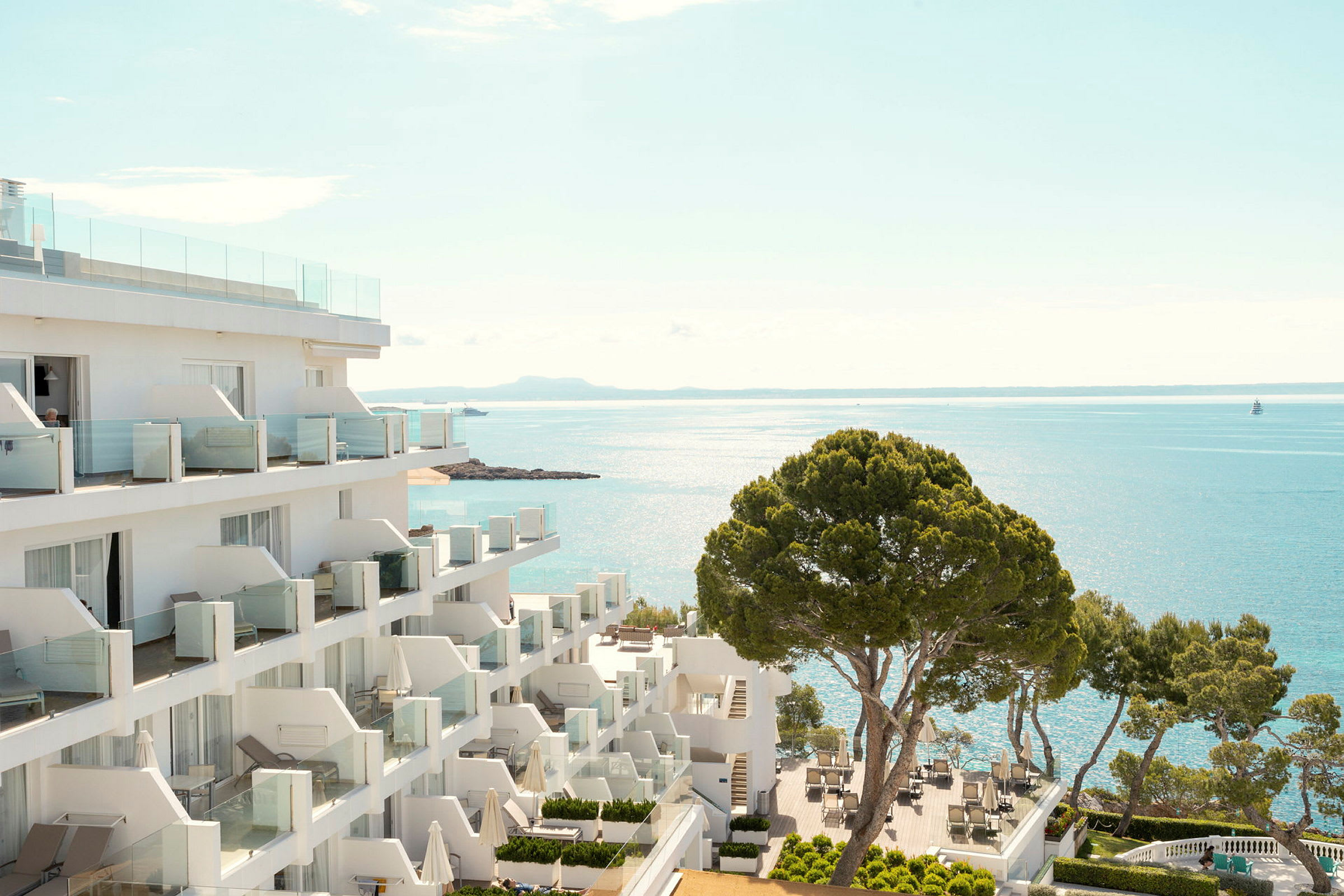 Hotelbygning ved kysten med udsigt over havet og en terrasse med borde og stole samt træer. (AI-genereret tekst)