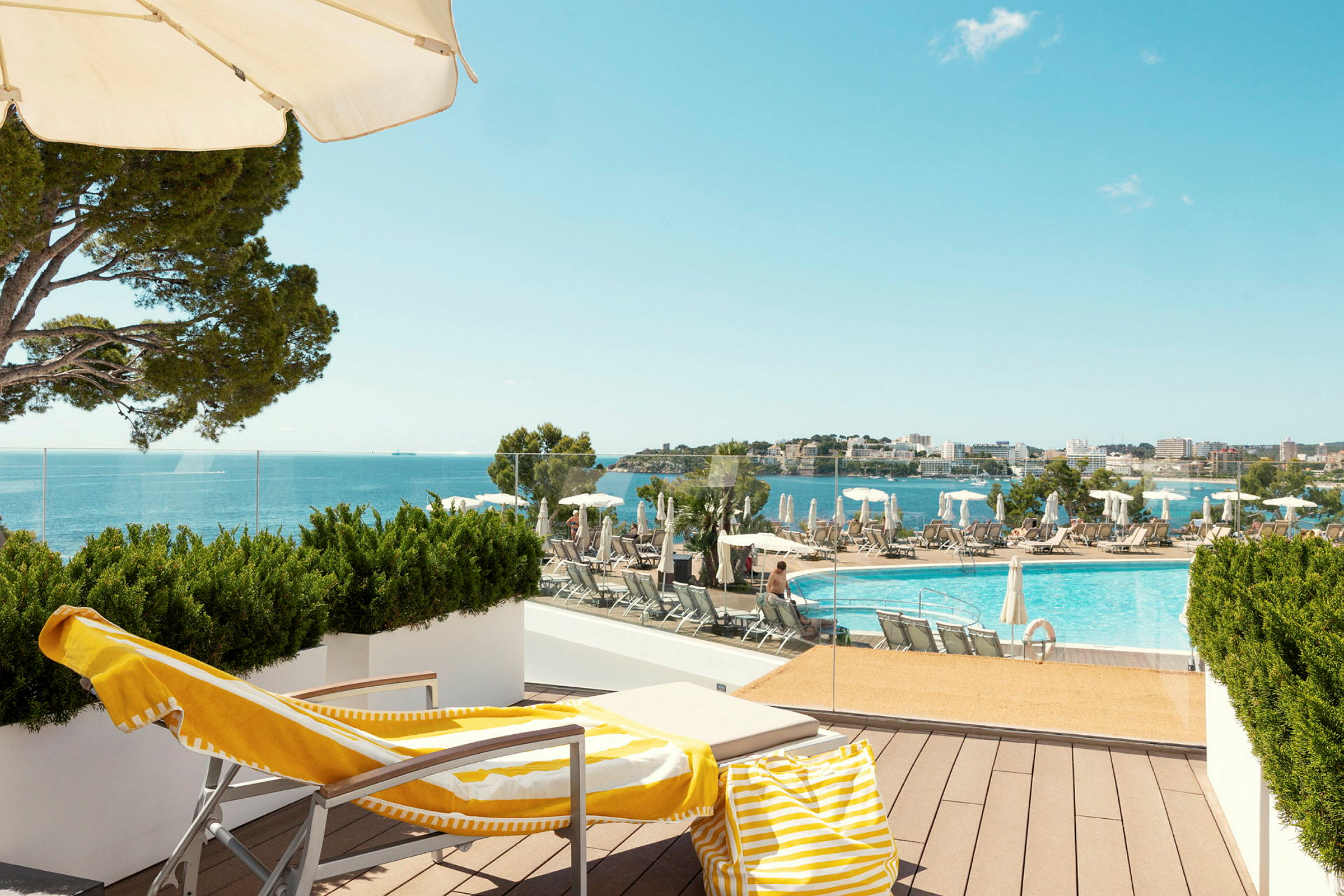 En liggestol med en gul-hvid håndklæde og strandtaske står på en terrasse med udsigt over en pool og havet. (AI-genereret tekst)