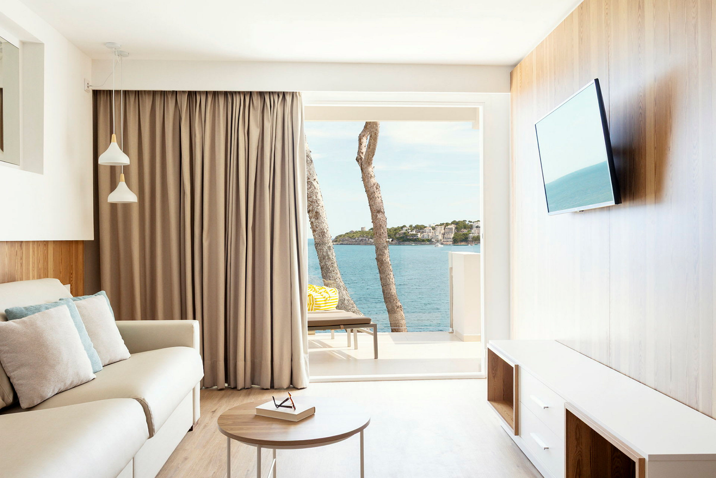 En lys stue med en sofa, bord og fjernsyn. Vinduesudsigten viser havet og nogle træer. (AI-genereret tekst)