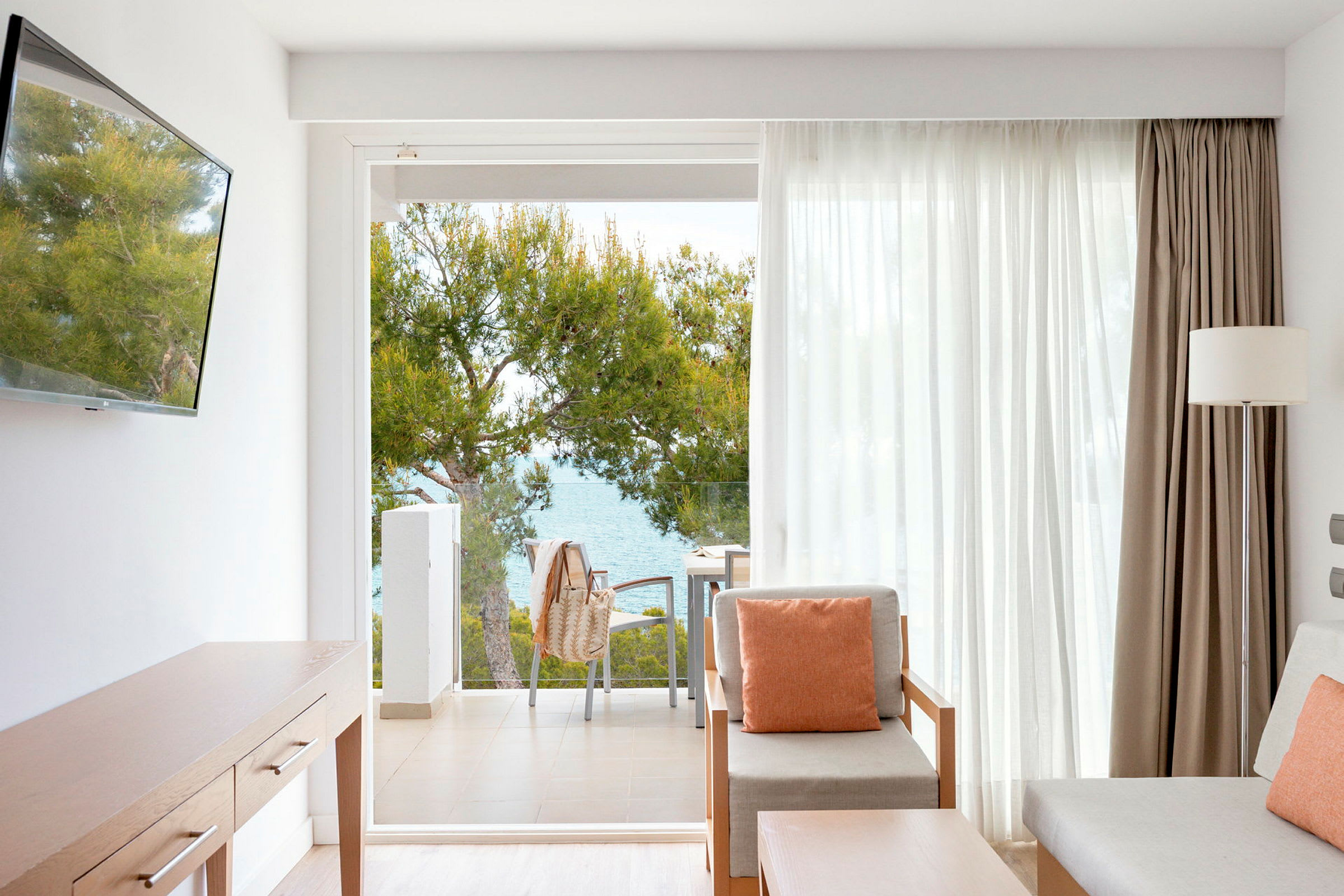 Et hotelværelse med siddeområde og udsigt til en balkon, med træer og hav i baggrunden. (AI-genereret tekst)