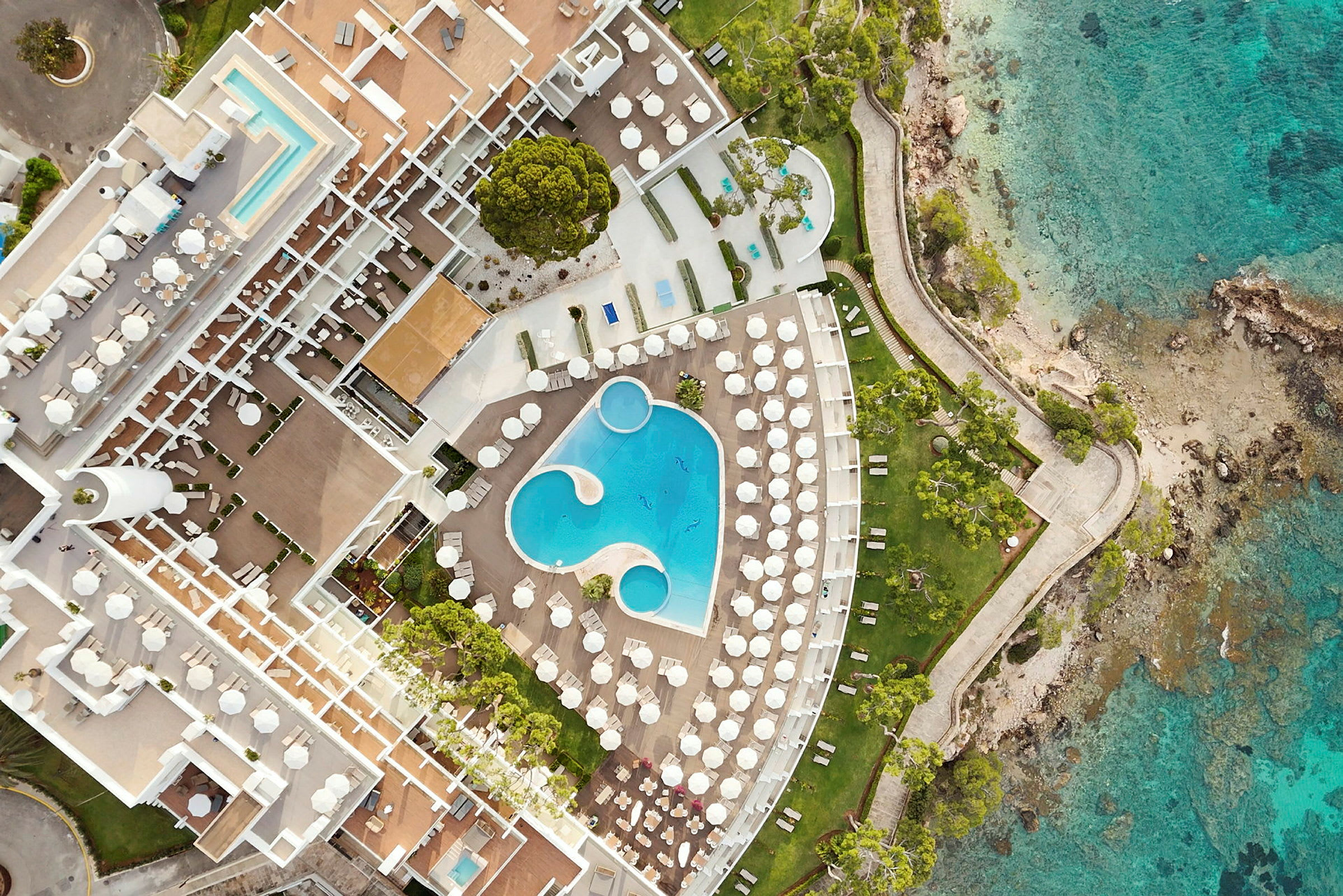 Luftfoto af et hotelområde med pools, liggestole og kystlinje. (AI-genereret tekst)