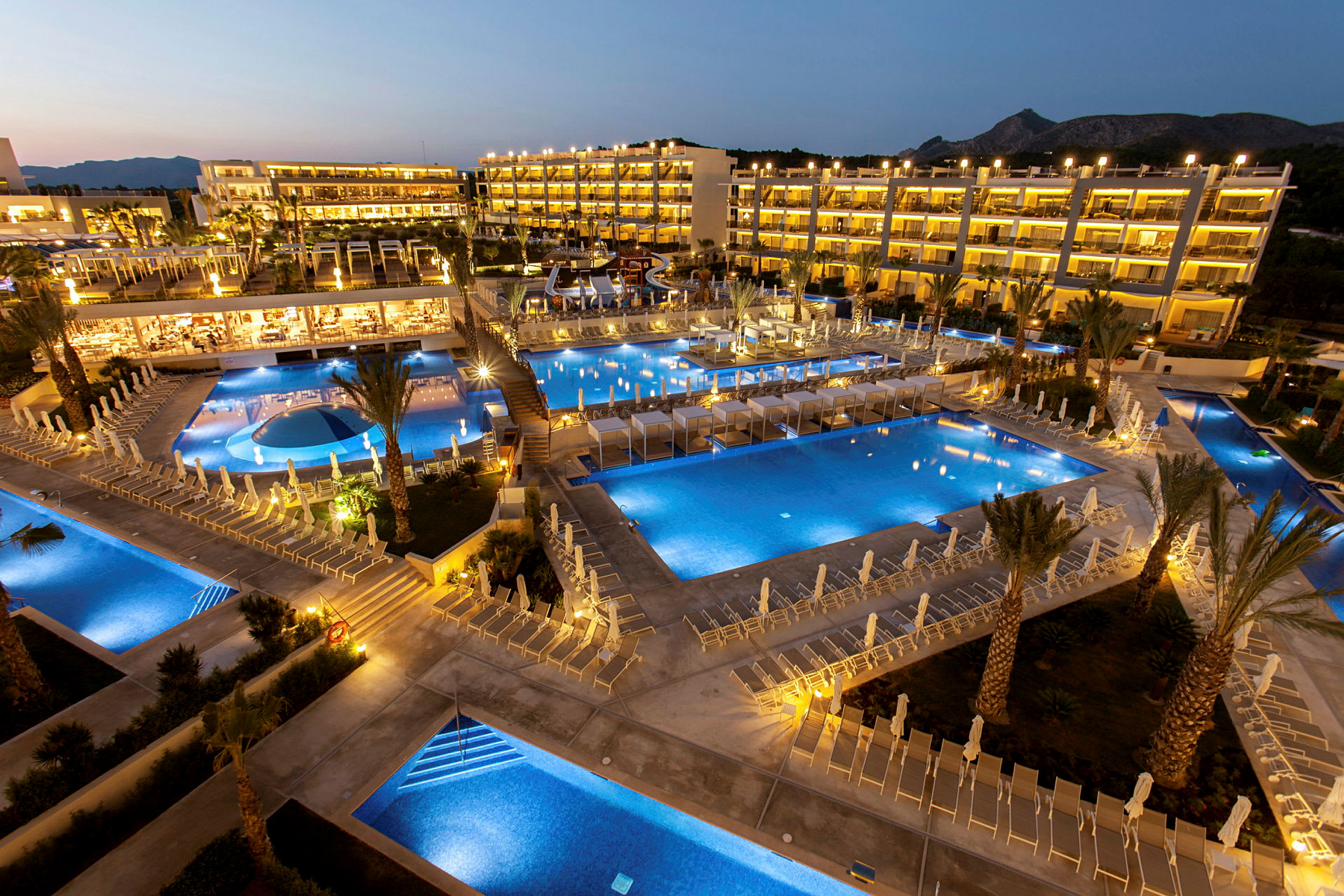 Et hotelområde om aftenen med flere oplyste swimmingpools og liggestole. (AI-genereret tekst)