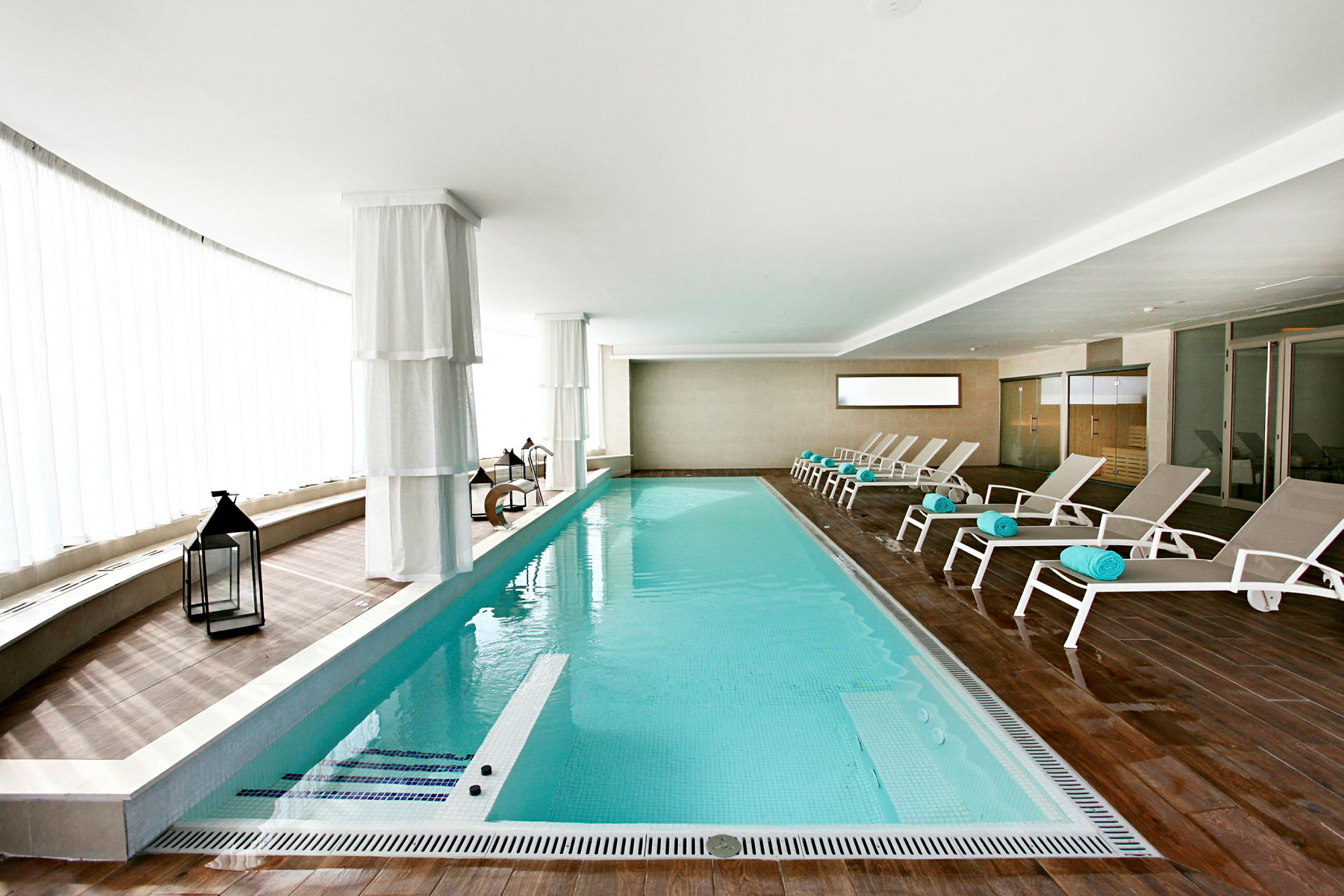 Indendørs swimmingpool med liggestole og hvide gardiner. (AI-genereret tekst)