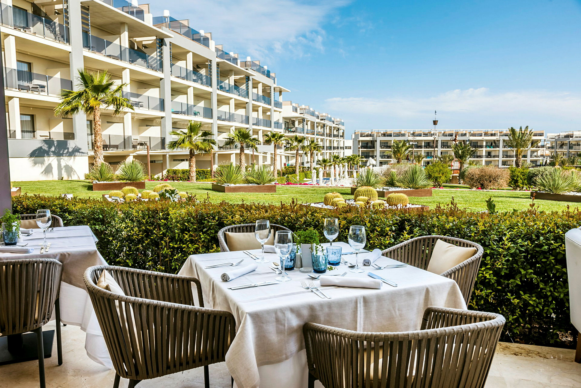 Billede af en udendørs restaurant med dækkede borde, omgivet af en have og moderne boliger i baggrunden. (AI-genereret tekst)