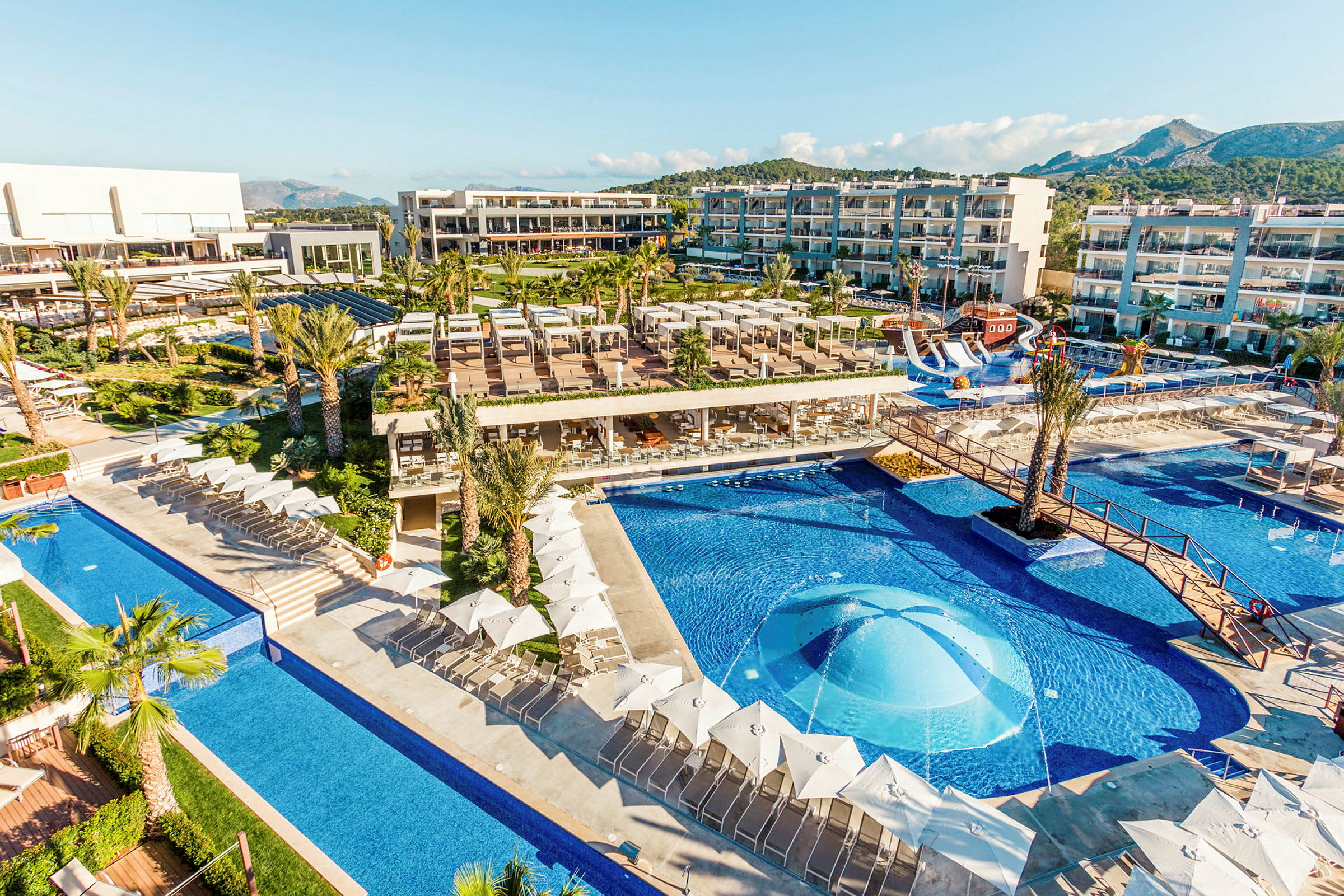 En udsigt over et hotelområde med pools, liggestole og bygninger. (AI-genereret tekst)