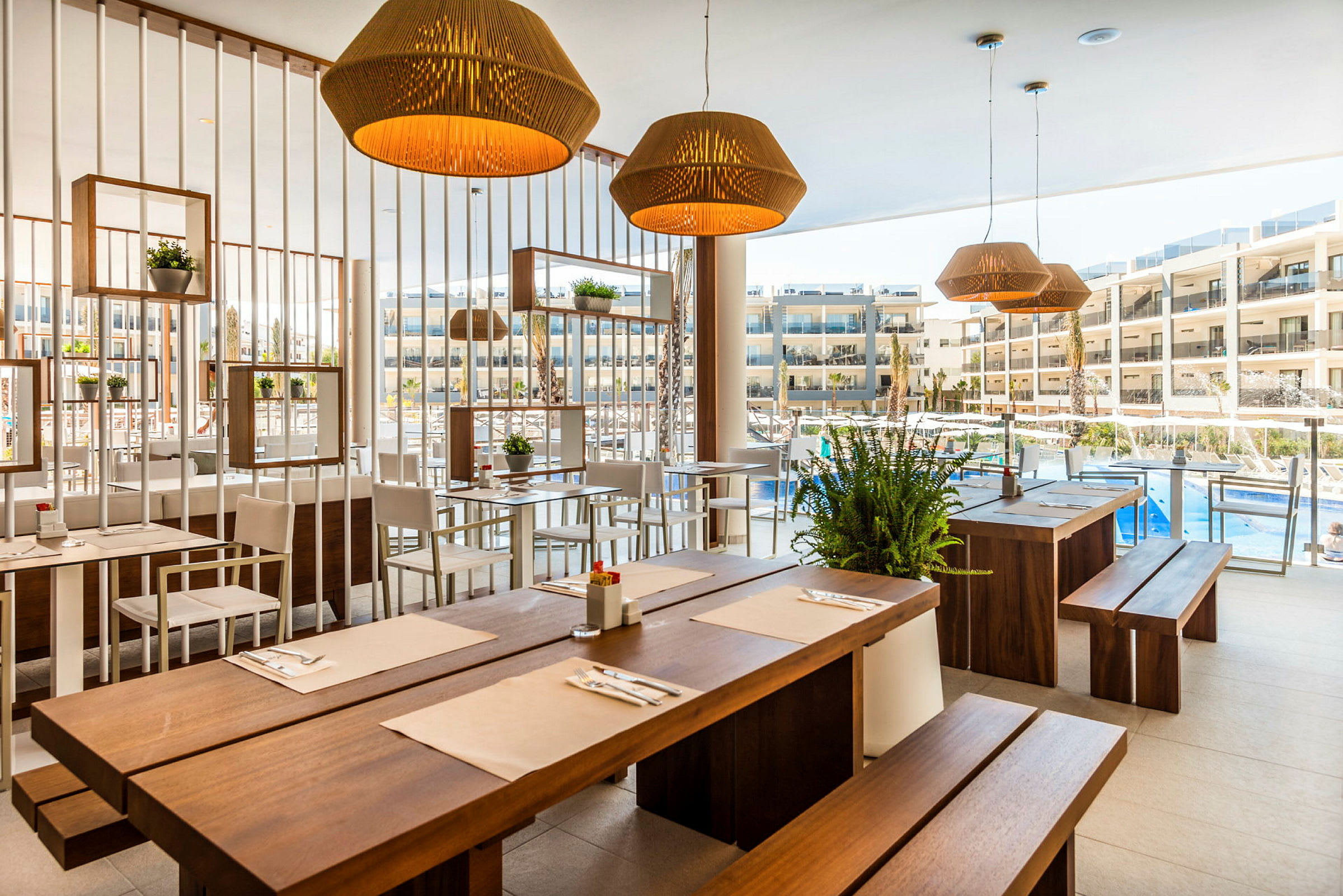 Restaurant med træborde og lyse stole, dekoreret med planter og lamper, med udsigt til en pool og hotelbygninger. (AI-genereret tekst)
