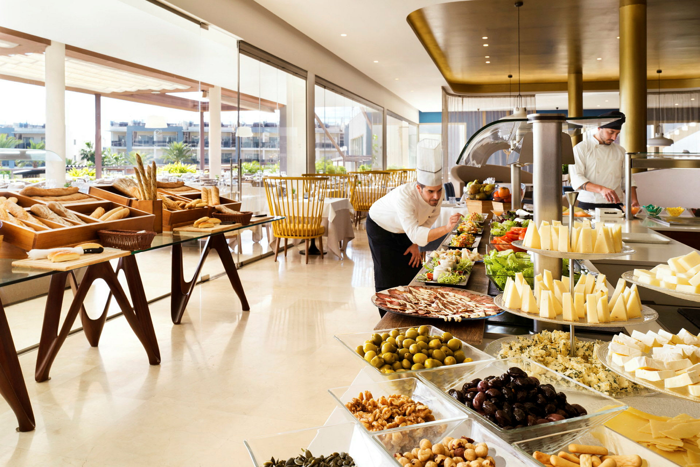 Billede af en lys restaurant med store vinduer og en buffet med forskellige retter, herunder brød, ost, salater og oliven. (AI-genereret tekst)