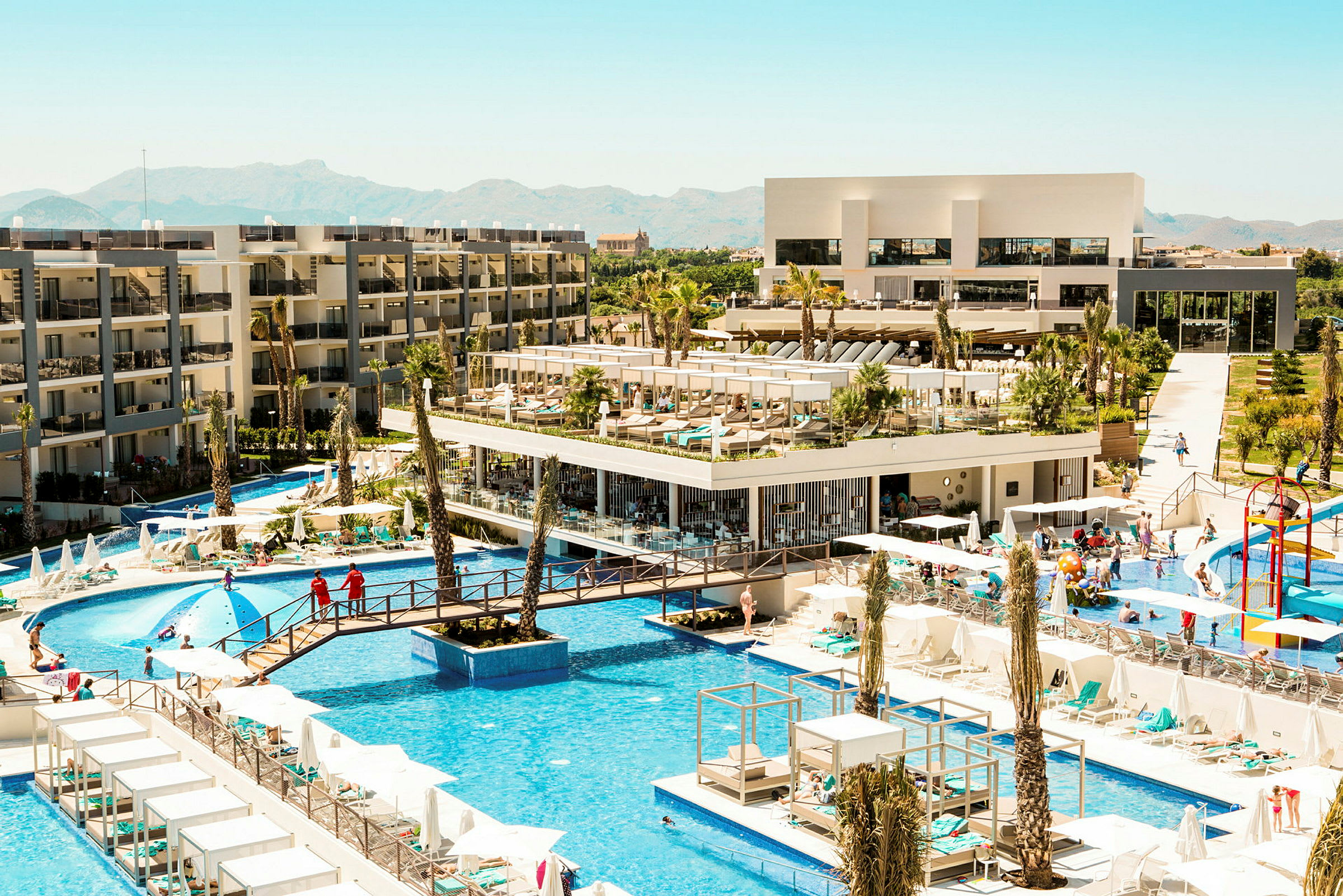 Udsigt over hotelområde med pools, bygninger og liggestole. (AI-genereret tekst)