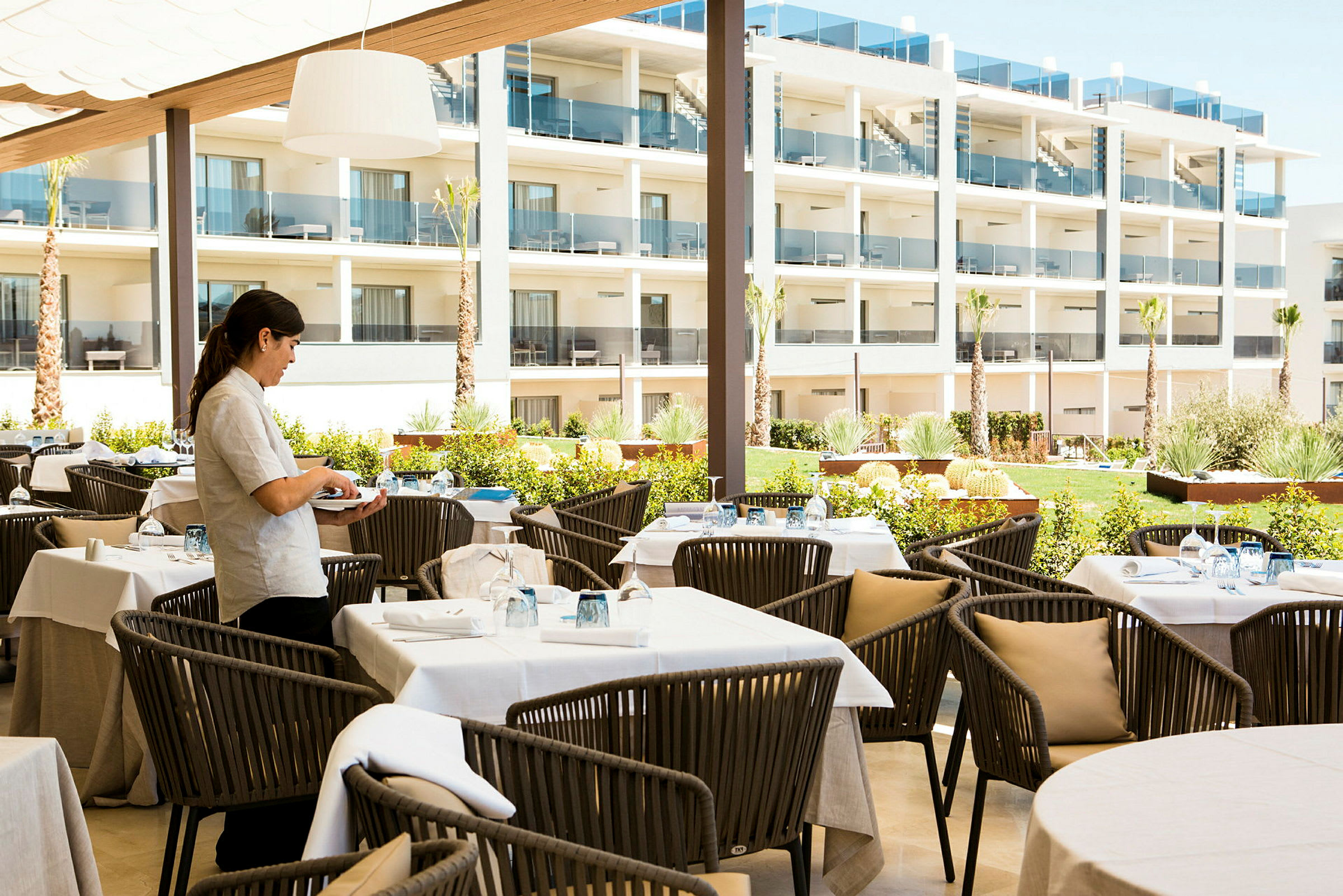 Udendørs restaurantområde med møbler og hvidklædte borde med en fleretagers bygning i baggrunden. (AI-genereret tekst)