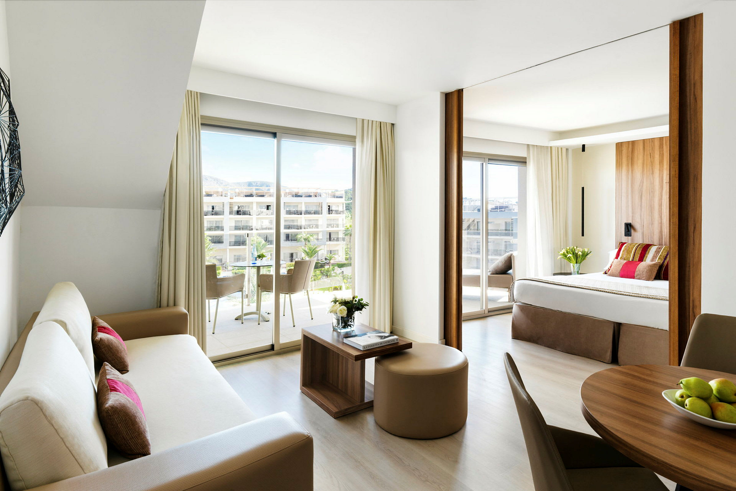 Et moderne hotelværelse med åben planløsning, inkluderer en stue med sofa og et soveværelse med dobbeltseng samt store vinduer med udsigt mod en terrasse. (AI-genereret tekst)