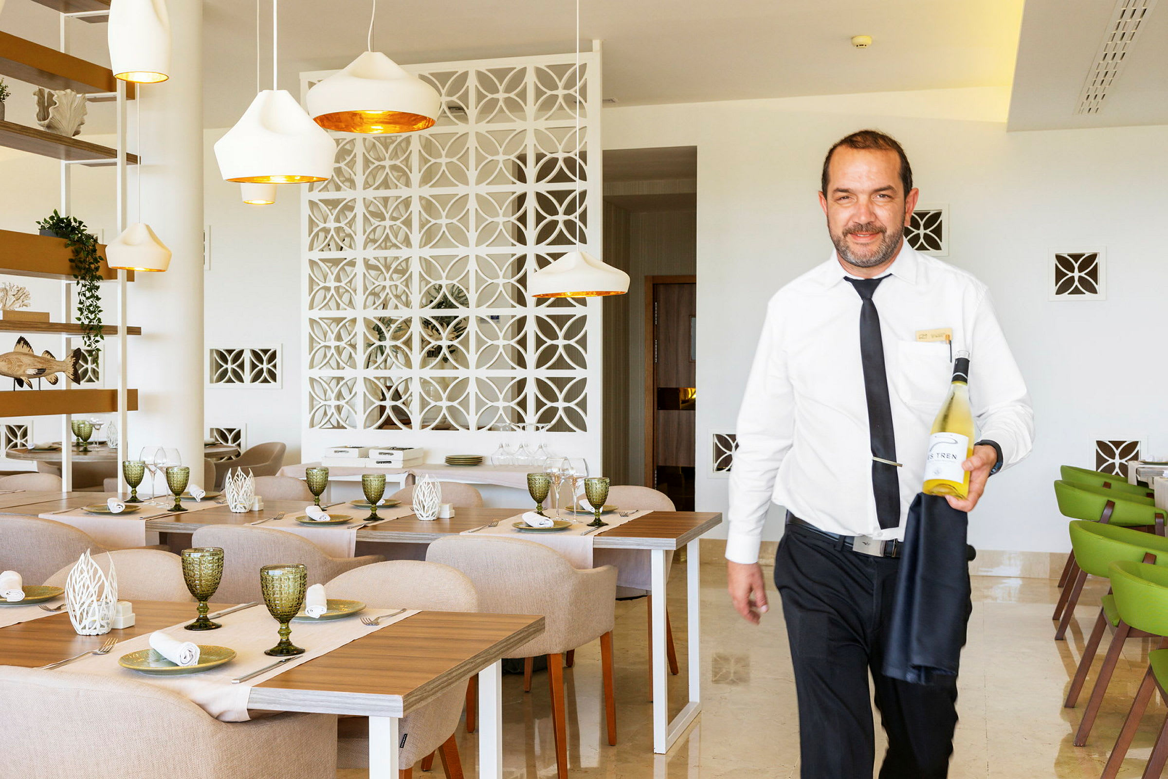 En tjener i hvid skjorte holder en flaske vin i en lys og moderne restaurant. (AI-genereret tekst)