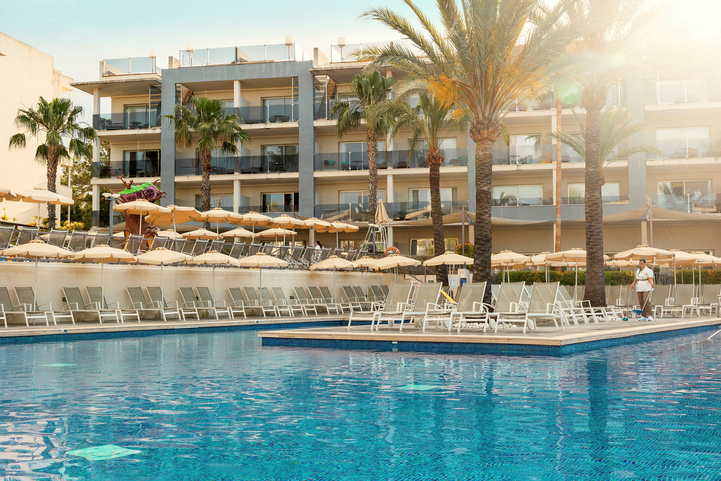 En udendørs pool omgivet af liggestole og parasoller foran et hotel med flere etager. (AI-genereret tekst)