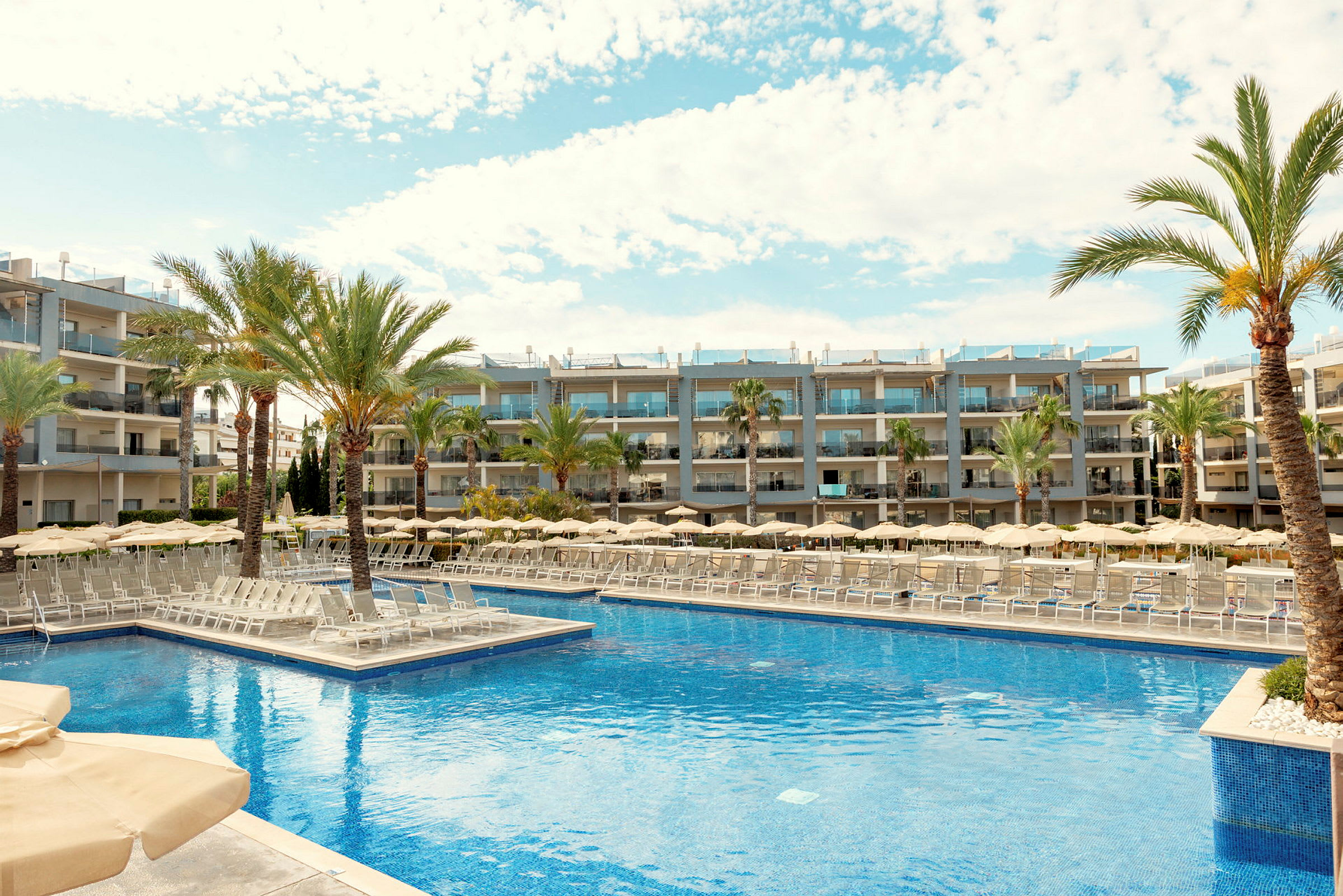 Et billede af et hotelområde med en stor pool omgivet af solvogne, parasoller og palmer. I baggrunden ses moderne hotelbygninger. (AI-genereret tekst)