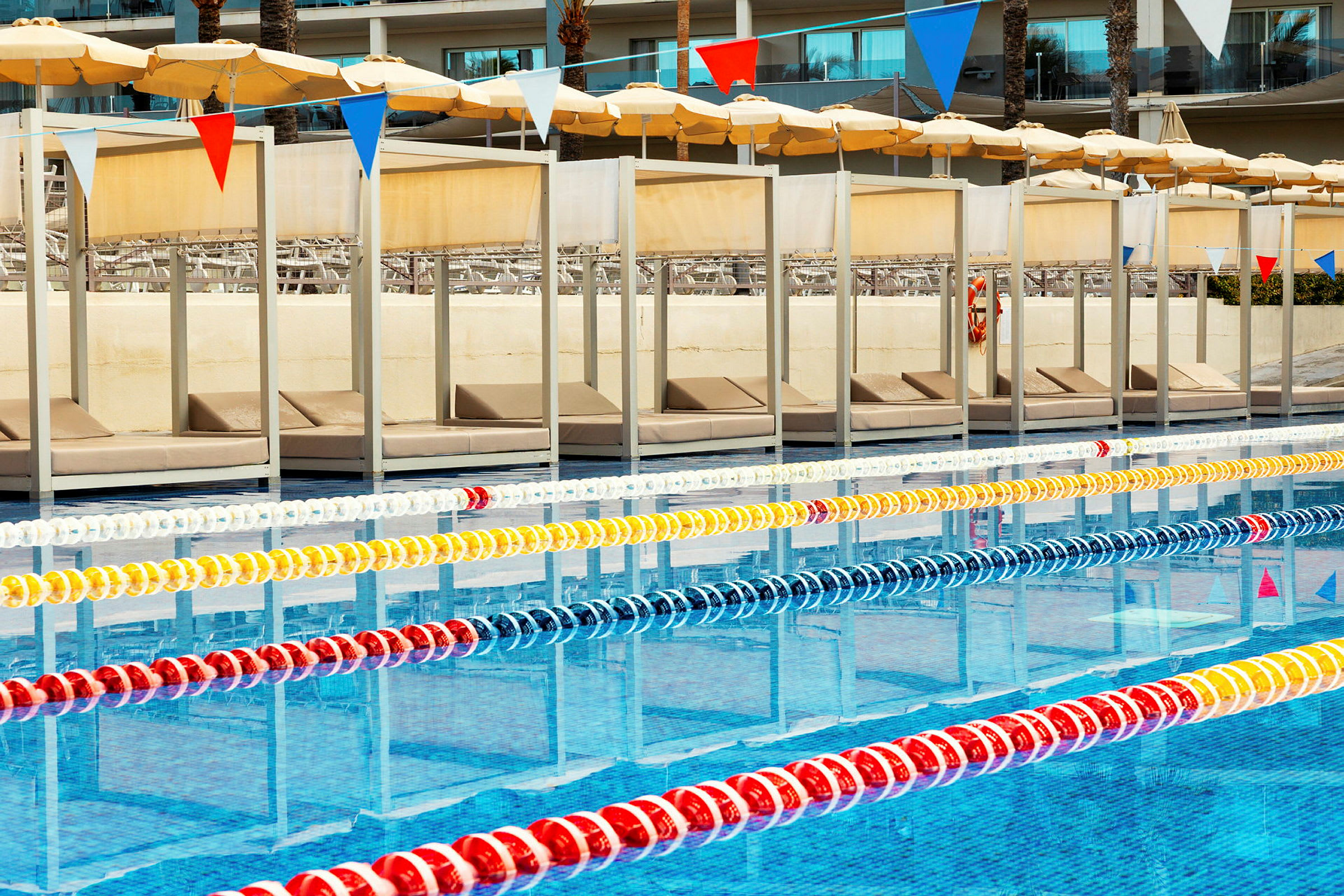 Swimmingpool med skillelinjer i hvidt, gult, rødt og blåt. I baggrunden ses liggestole og parasoller. (AI-genereret tekst)