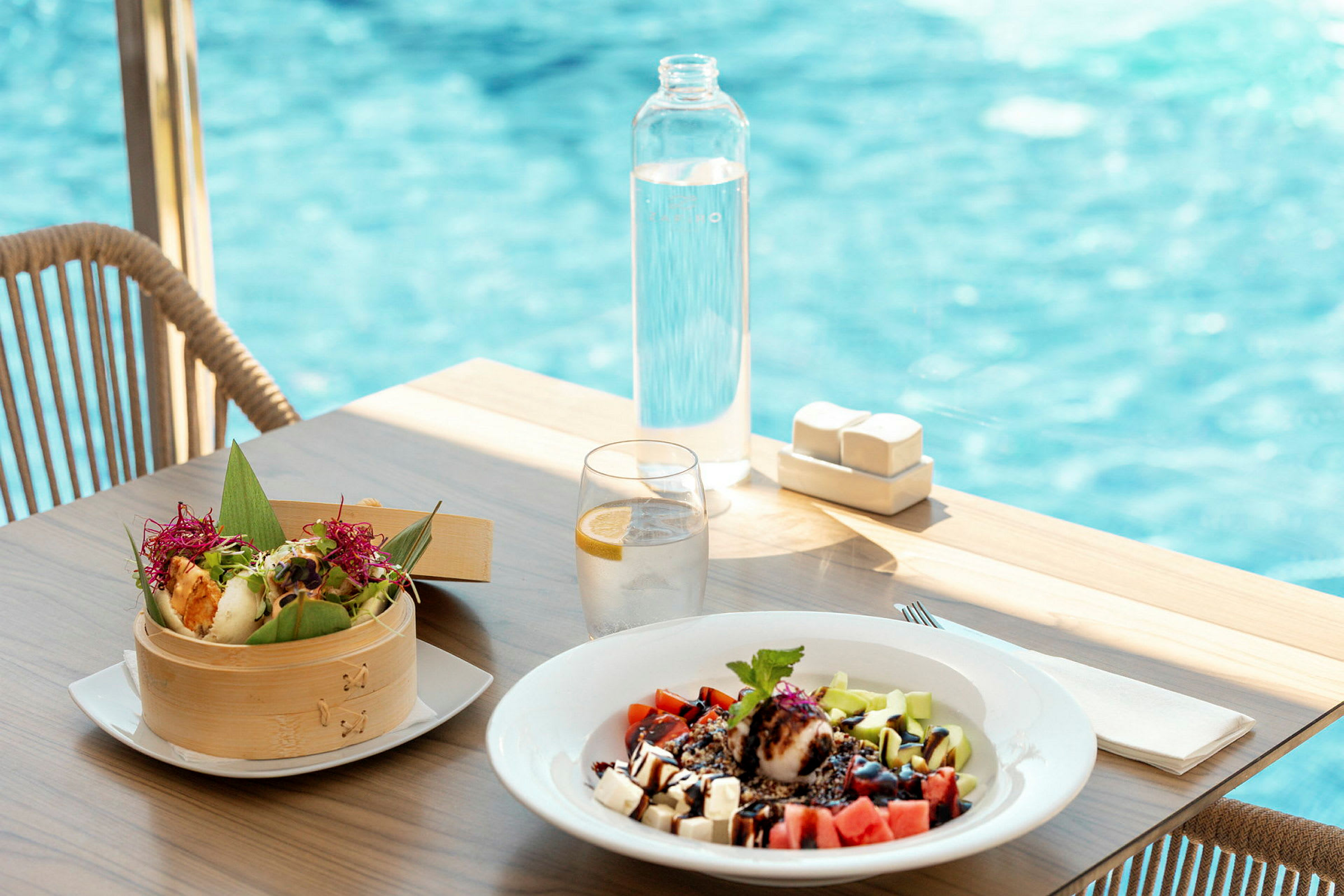 Et dækket bord med en skål med farverig mad, en tallerken med frugtsalat, et glas vand med citron og en flaske vand. En pool ses i baggrunden. (AI-genereret tekst)