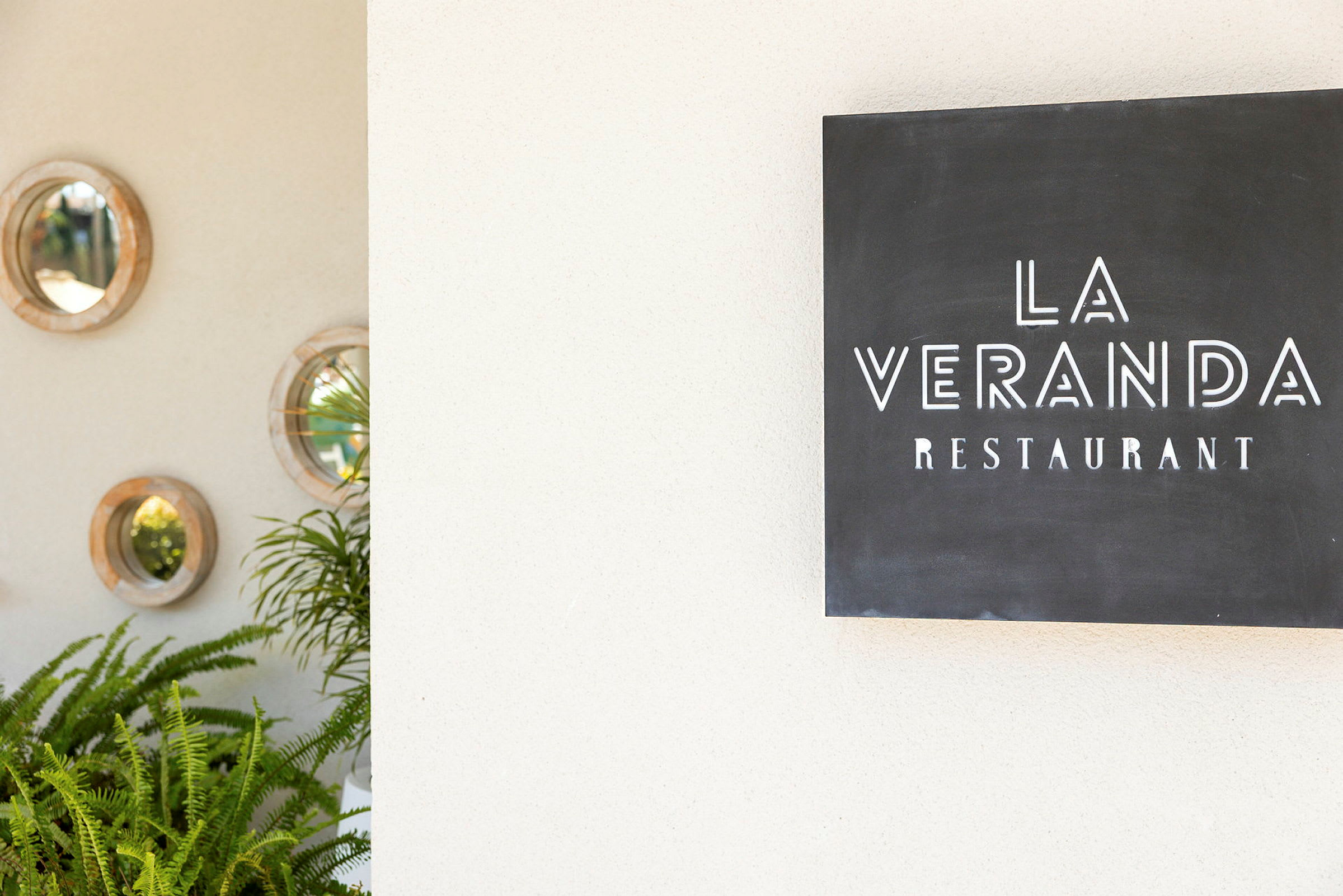 Et skilt med teksten "La Veranda Restaurant" ved siden af en væg dekoreret med runde spejle og grønne planter. (AI-genereret tekst)