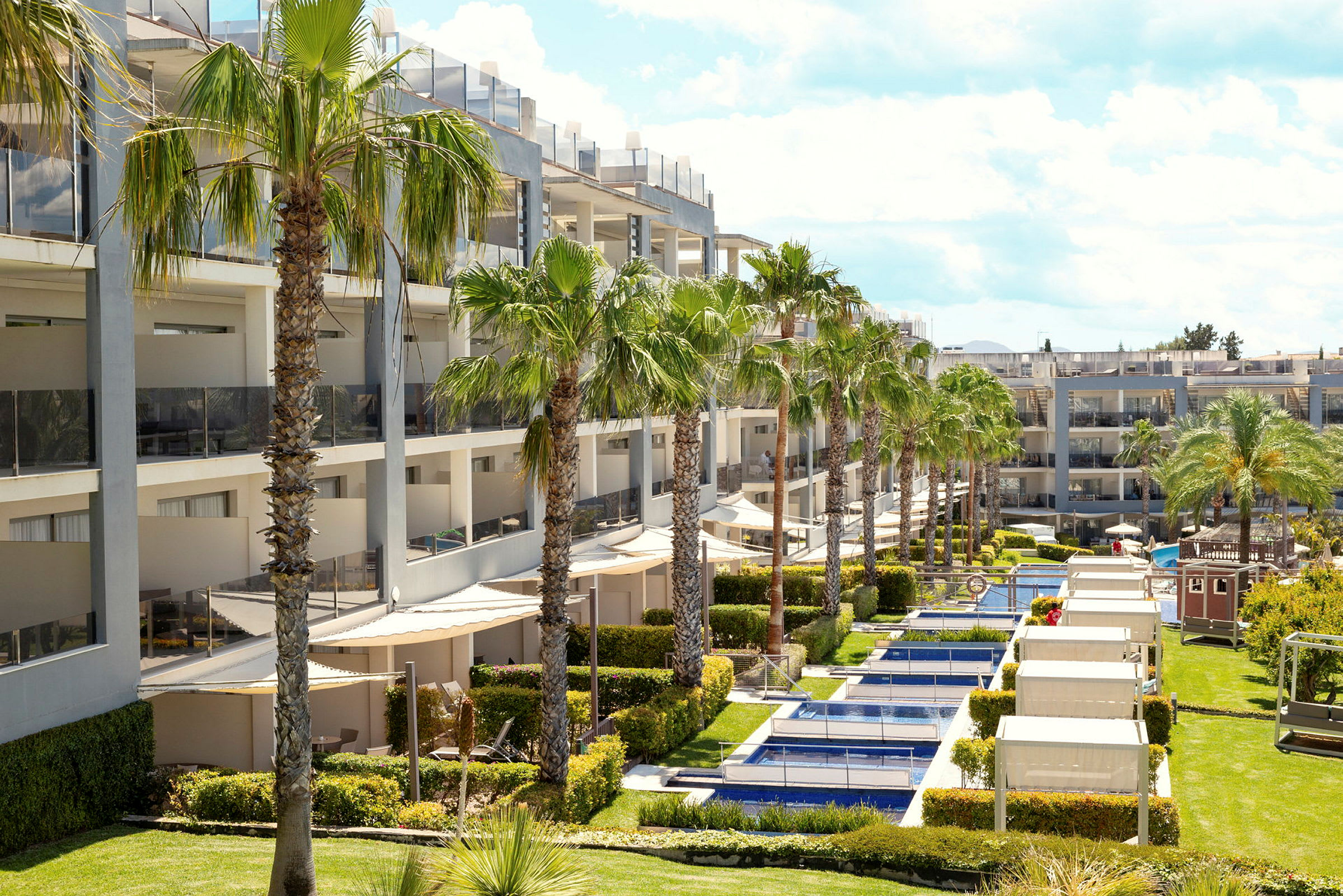 Et moderne hotel med flere etager og altaner. Foran bygningen ses pools og palmer. (AI-genereret tekst)
