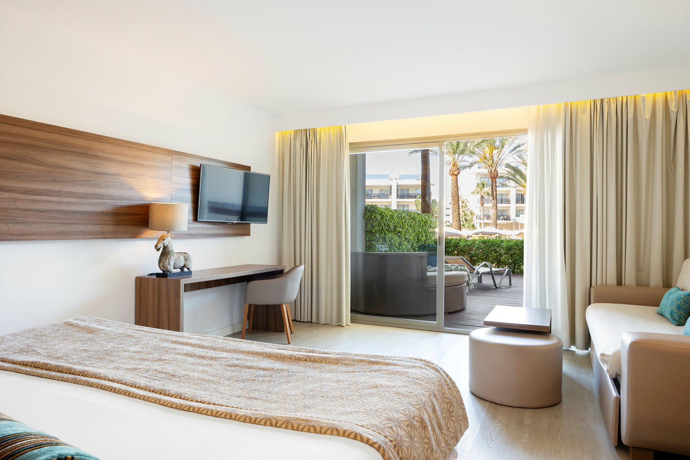 Moderne hotelværelse med en seng, sofa, skrivebord og et tv på væggen. Glasdør ud til en balkon med udsigt til en have. (AI-genereret tekst)