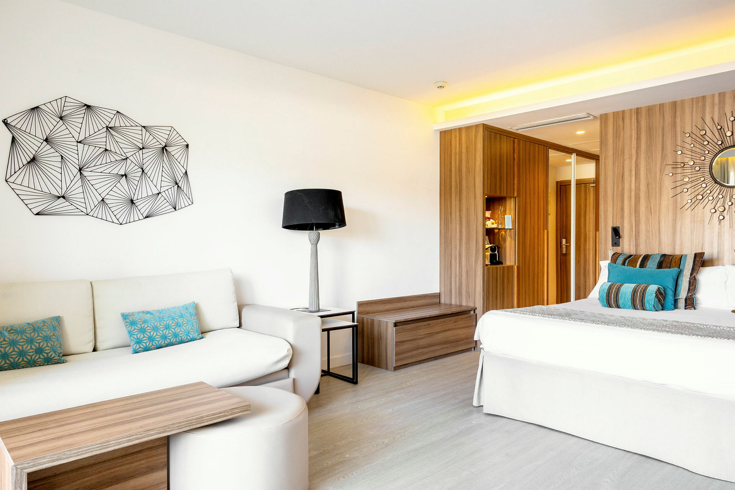 Et moderne hotelværelse med en sofa, farverige puder, et sofabord, en seng med dekorative puder og en træpanel bagved. På væggen er der kunst og en lille rundt spejldekoration. (AI-genereret tekst)