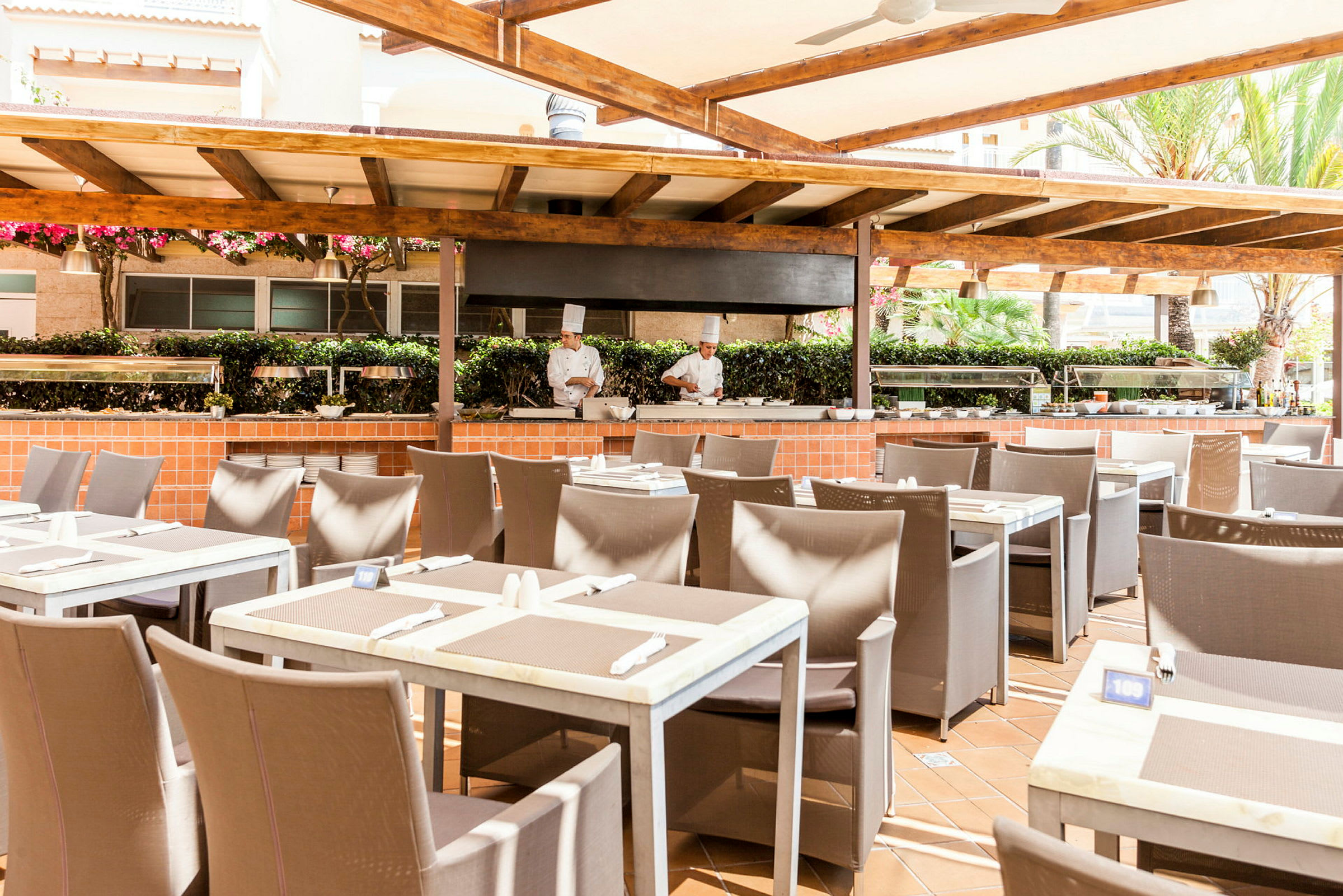 Udendørsrestaurant med dækkede borde og madlavningsstation under en træpergola. (AI-genereret tekst)