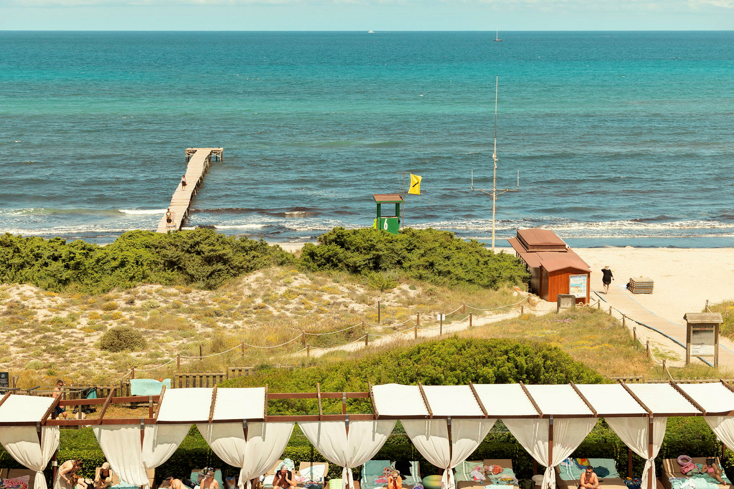 En strand med en bro, der strækker sig ud i havet, sandklitter med vegetation i forgrunden og personer i et afslapnings- eller solområde. (AI-genereret tekst)