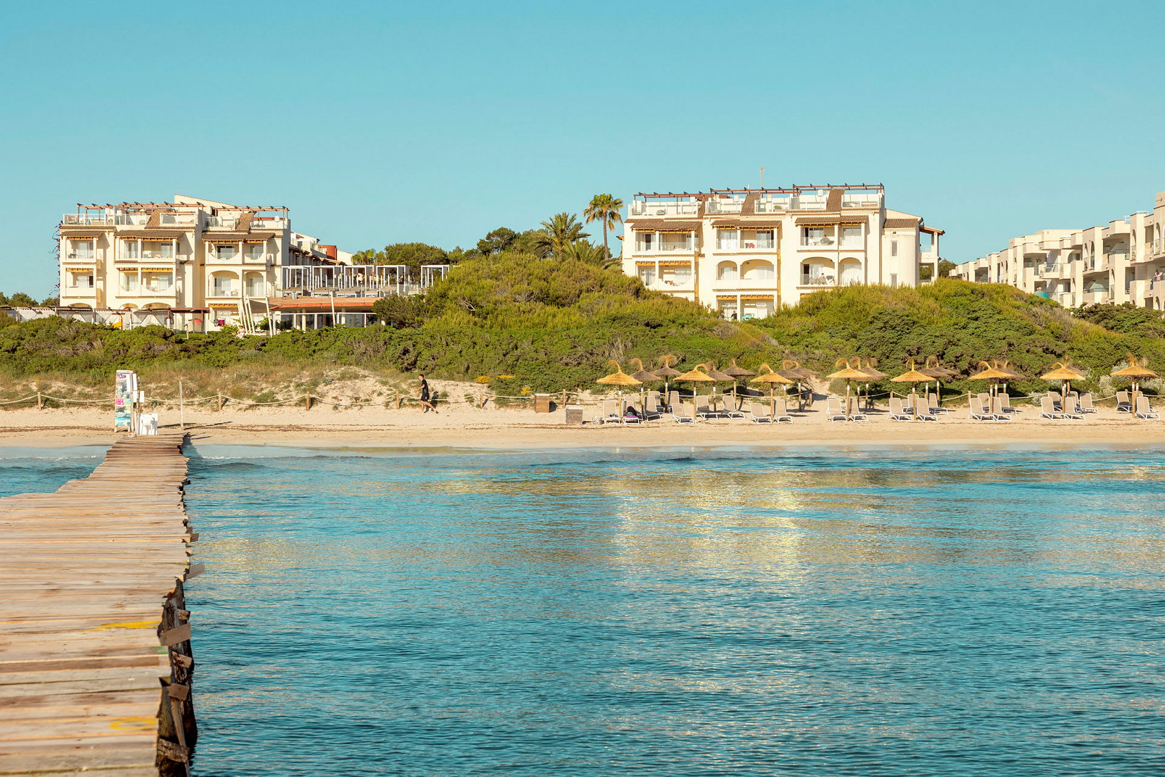 En strandpromenade fører til en sandstrand med parasoller og liggestole. I baggrunden er der hotelbygninger og beplantning. (AI-genereret tekst)