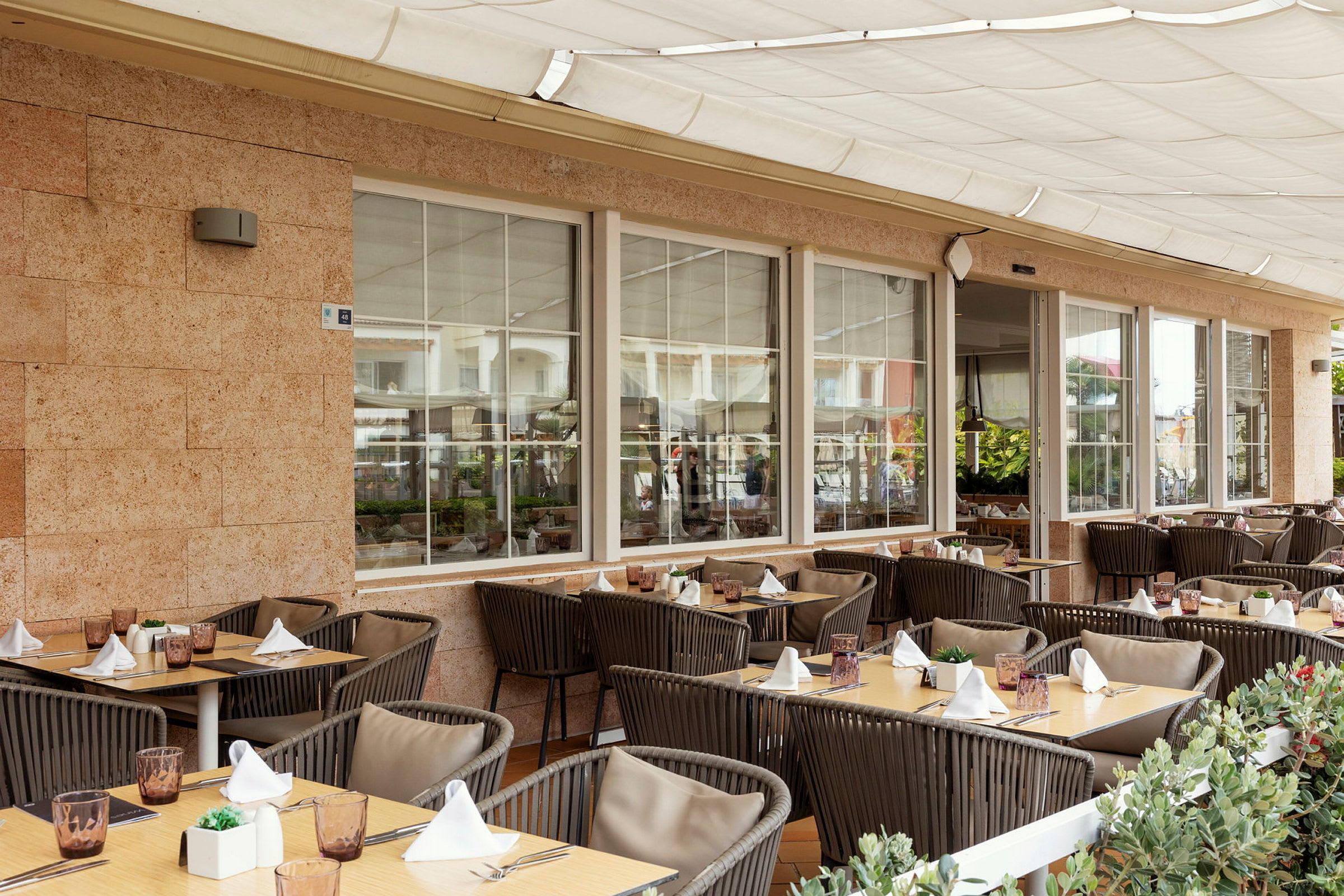 En udendørs terrasse ved en restaurant med dækkede borde og stole, omgivet af grønne planter. (AI-genereret tekst)