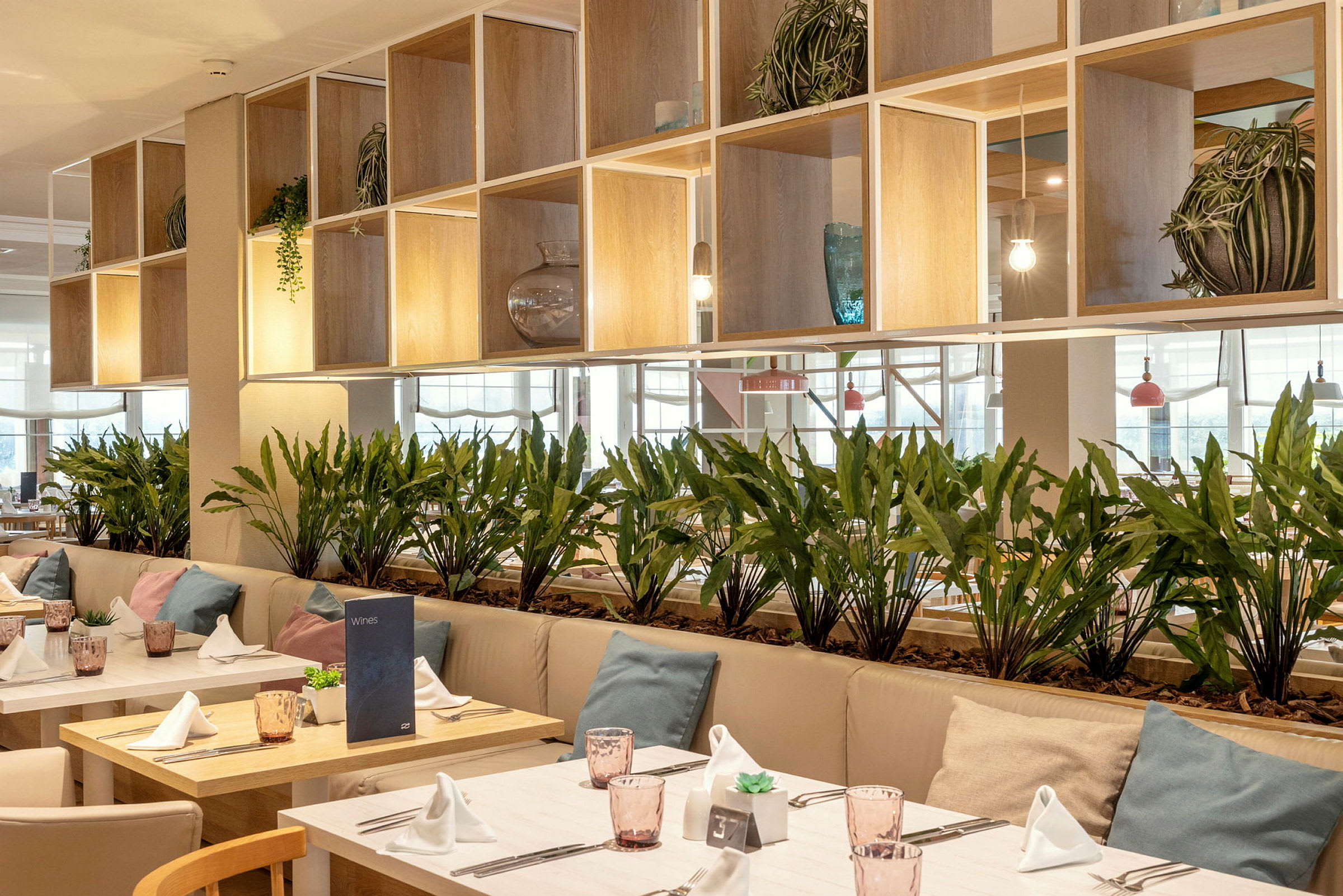 Et moderne indrettet restaurantområde med borde dekoreret med hvide servietter, glas og puder på sofaerne. Grønne planter placeret som skillevægge. (AI-genereret tekst)