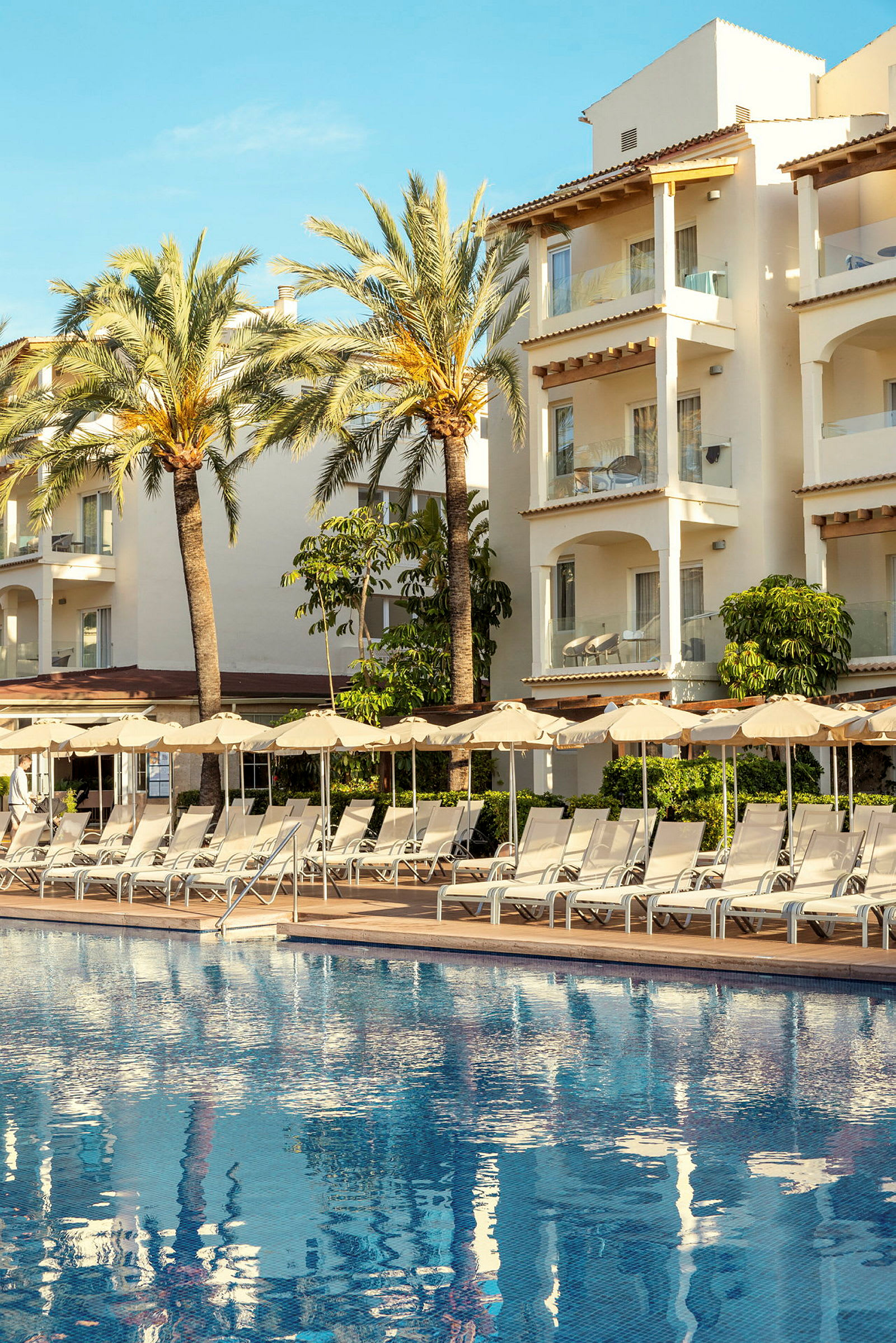 Billedet viser en udendørs pool med liggestole og parasoller foran en hotelbygning med altaner. (AI-genereret tekst)