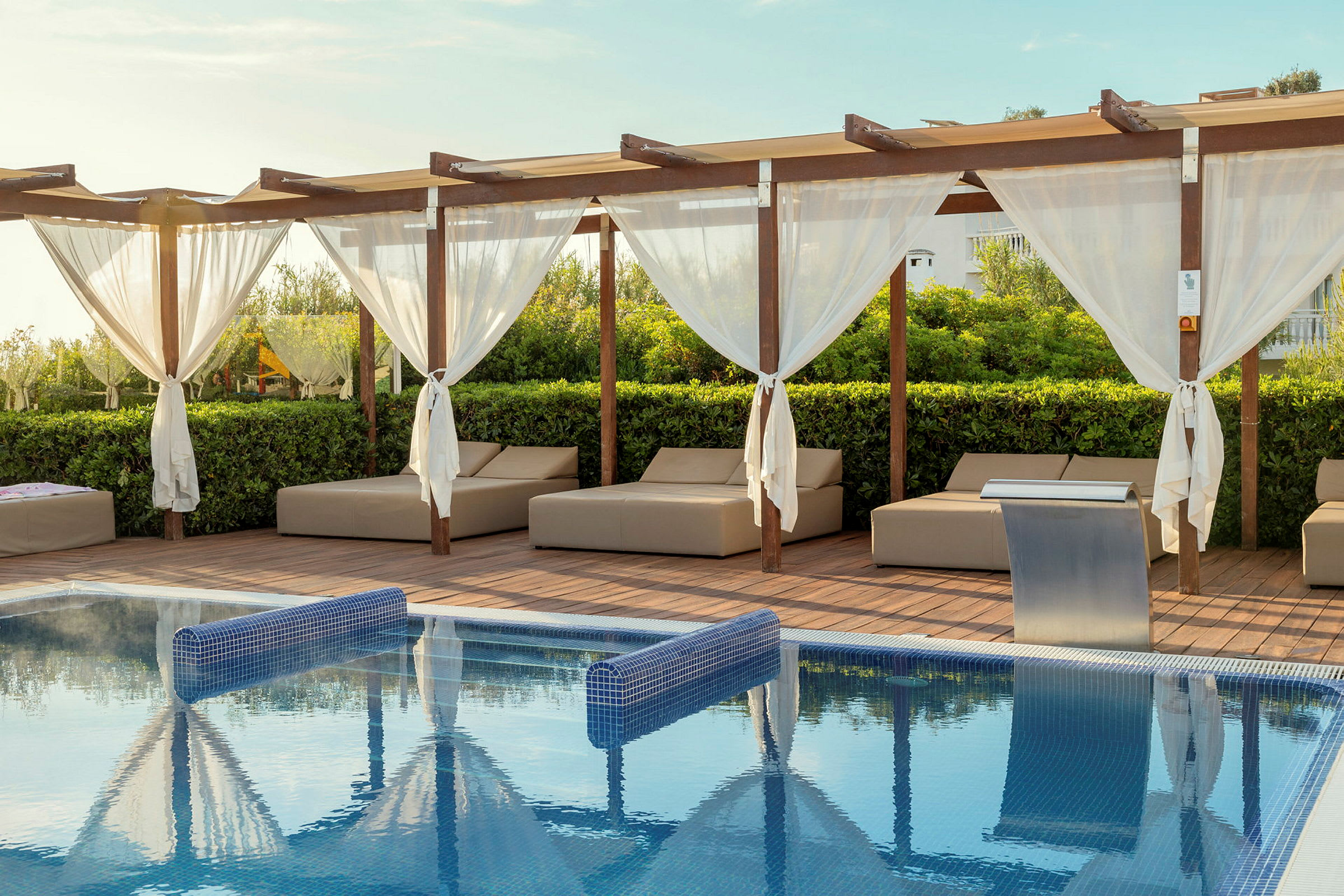 En pool med blåt vand, omgivet af liggestole og en træterrasse med pergola og hvide gardiner. (AI-genereret tekst)