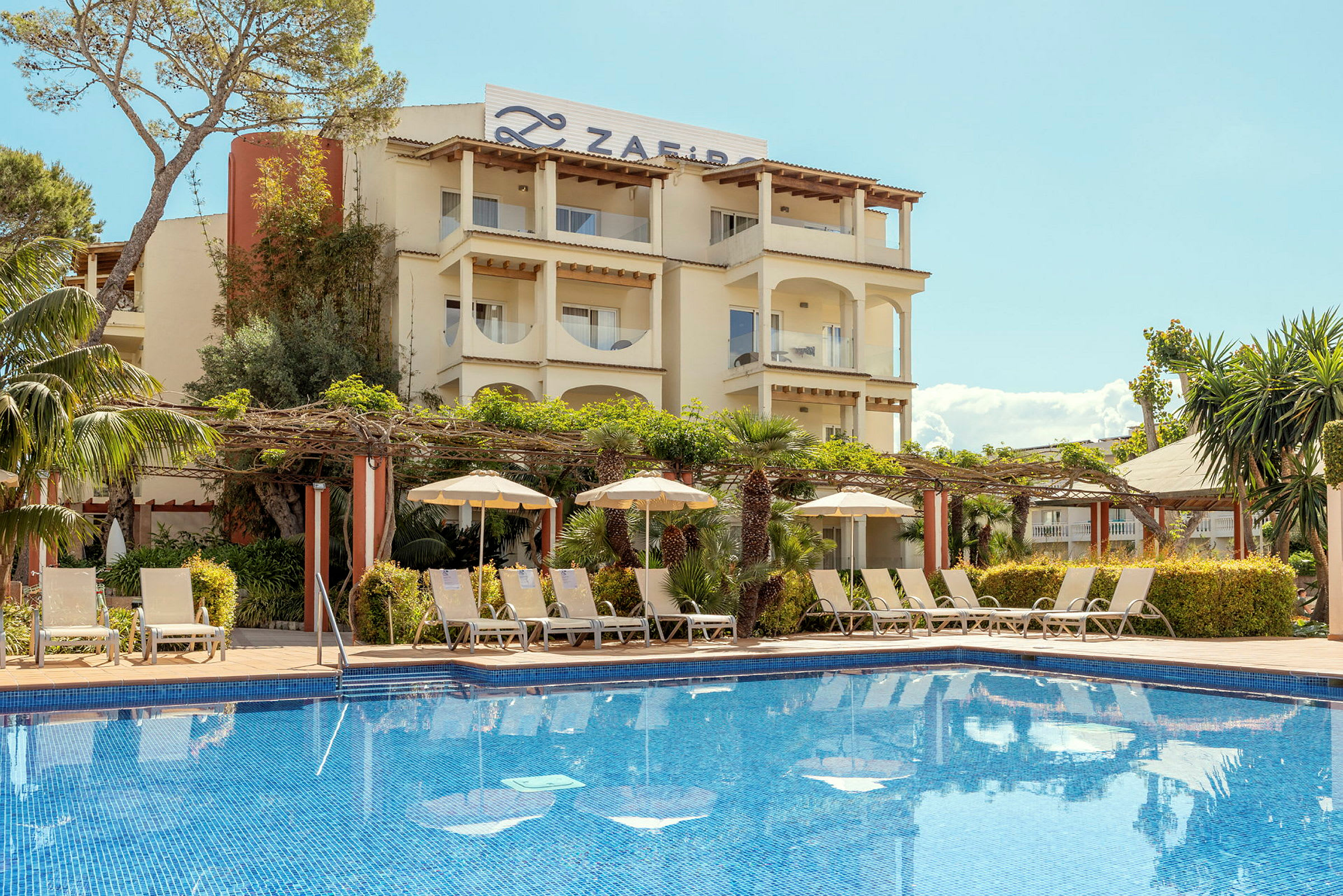 Et hotel med en swimmingpool i forgrunden, omgivet af liggestole og parasoller. Bygningen er omgivet af grønne områder og har træbjælker og søjler på altanerne. (AI-genereret tekst)