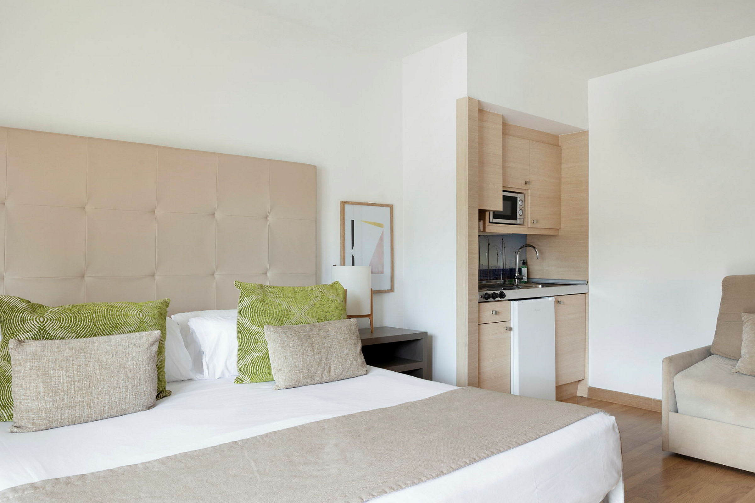 Et hotelværelse med en dobbeltseng, grønne og beige puder, en lille køkkenkrog med mikrobølgeovn og håndvask samt en beige lænestol. (AI-genereret tekst)