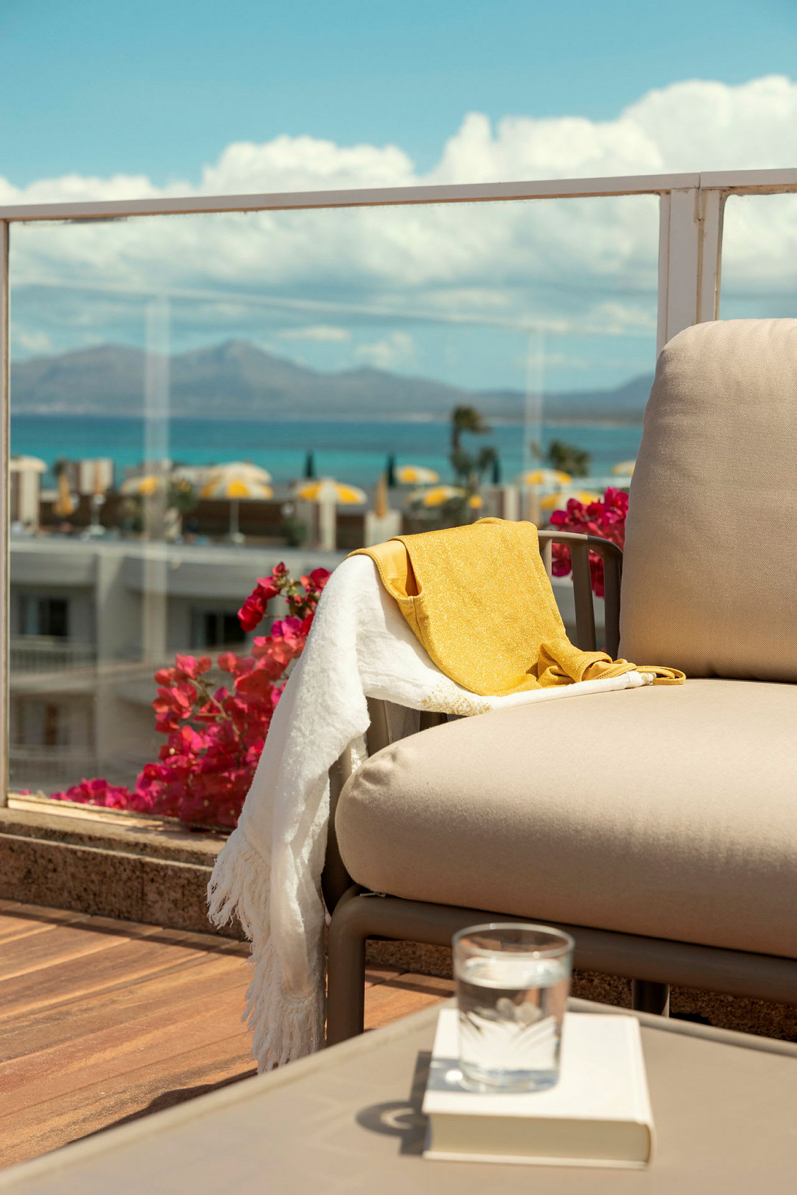 En beige stol på en terrasse med udsigt over havet og det omkringliggende landskab, med et gult og hvidt håndklæde hængende over armlænet. (AI-genereret tekst)