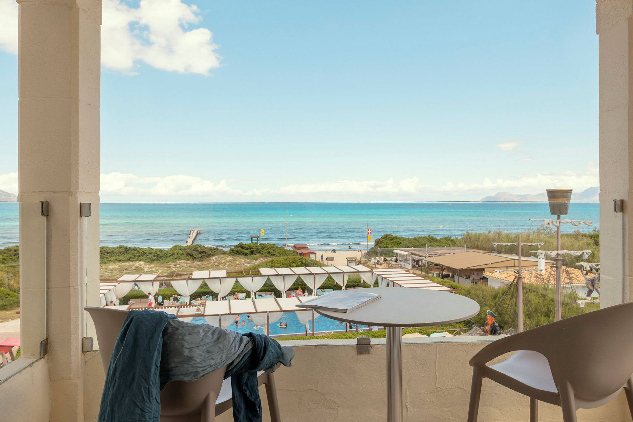 Udsigt fra en terrasse mod et poolområde og en strand med turkisblåt hav i baggrunden. (AI-genereret tekst)