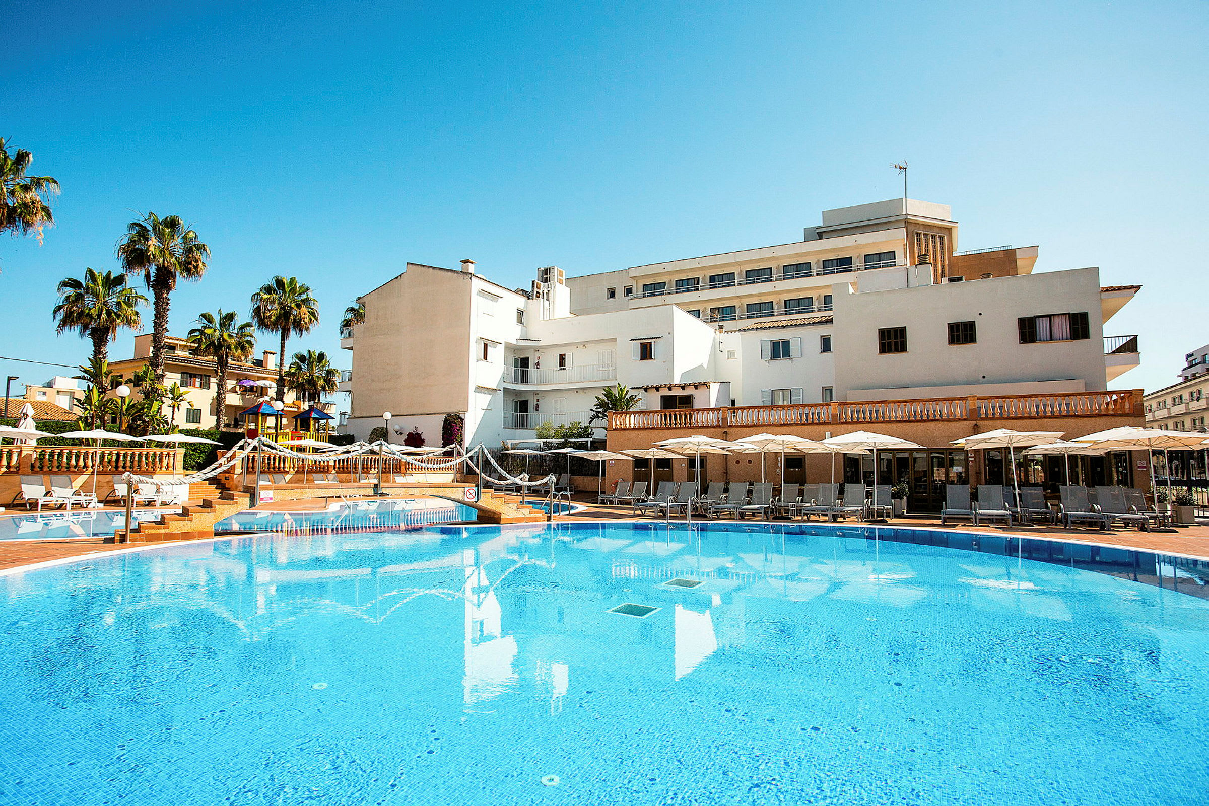Billede af hotel med pool, liggestole og parasoller, med palmer i baggrunden. (AI-genereret tekst)
