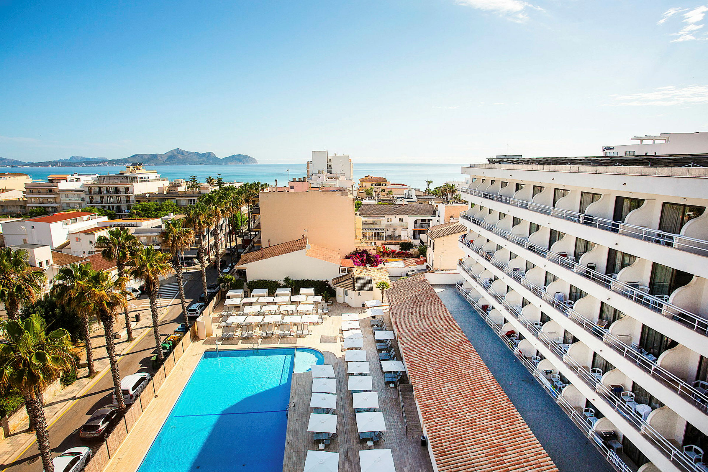 Udsigt fra oven af en hotelbygning, pool og omkringliggende område med havet i baggrunden. (AI-genereret tekst)