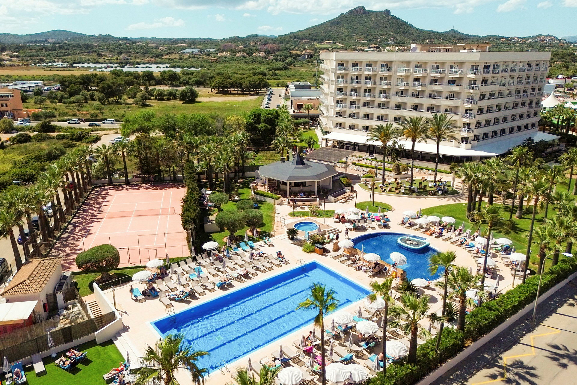 Hotelkompleks med swimmingpools, liggestole, tennisbane og have i forgrunden samt omkringliggende grønne områder og bjerge i baggrunden. (AI-genereret tekst)