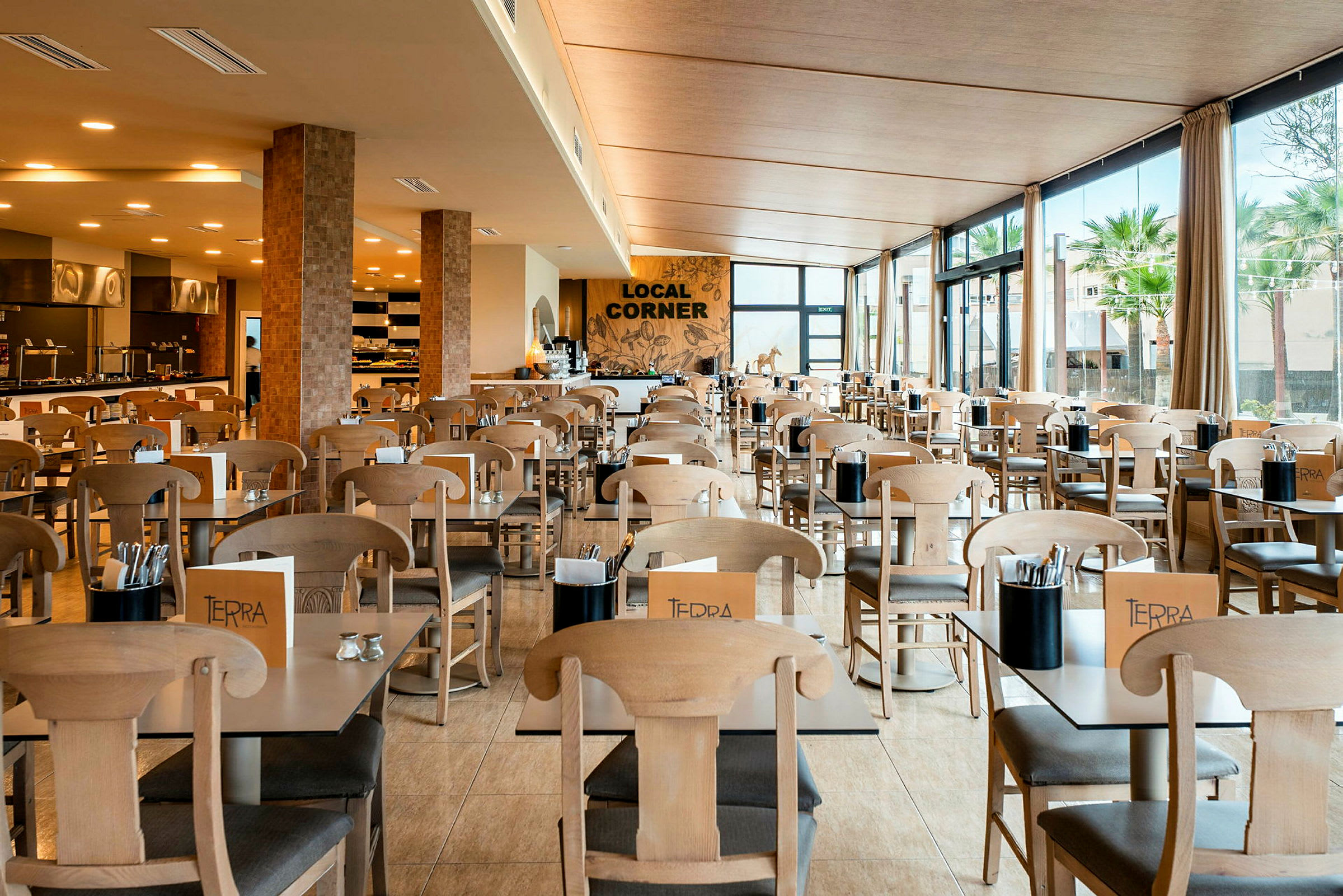 Et lyst og rummeligt restaurantområde med borde og træstole. Store vinduer med udsigt over grønne områder. (AI-genereret tekst)