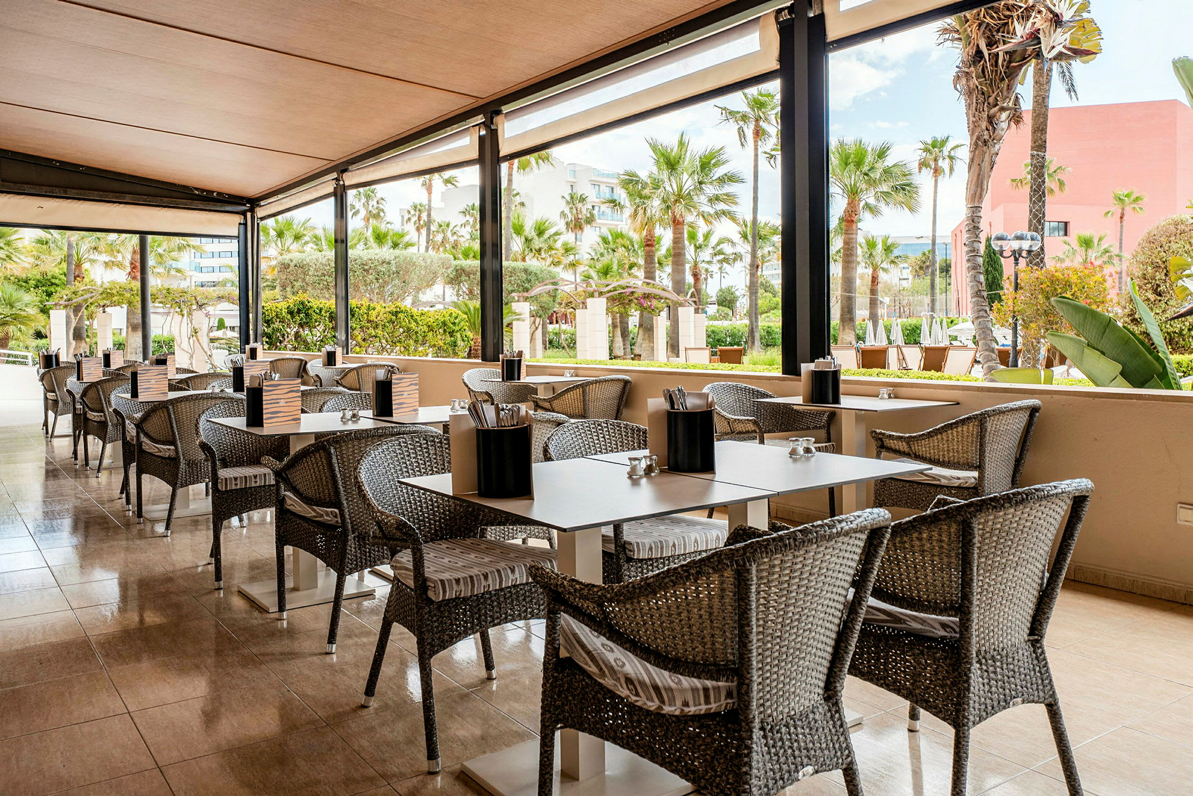 En udendørs restaurant med borde og stole på en terrasse med udsigt til en have med palmer. (AI-genereret tekst)