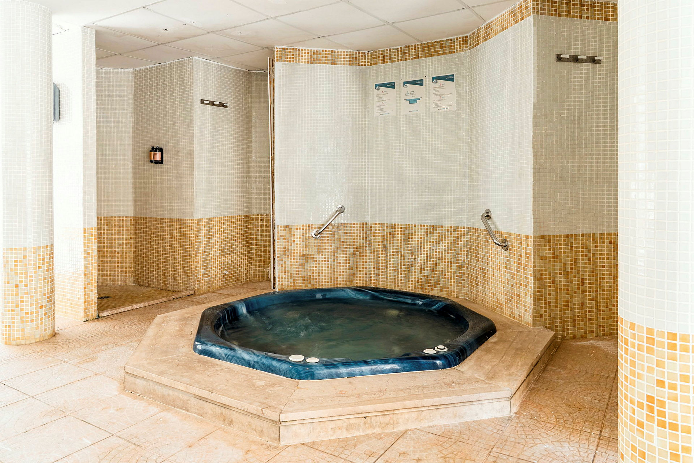Billedet viser et rum med en boblebad omgivet af mosaikdekorerede vægge. Boblebadet er blåt og integreret i gulvet. (AI-genereret tekst)