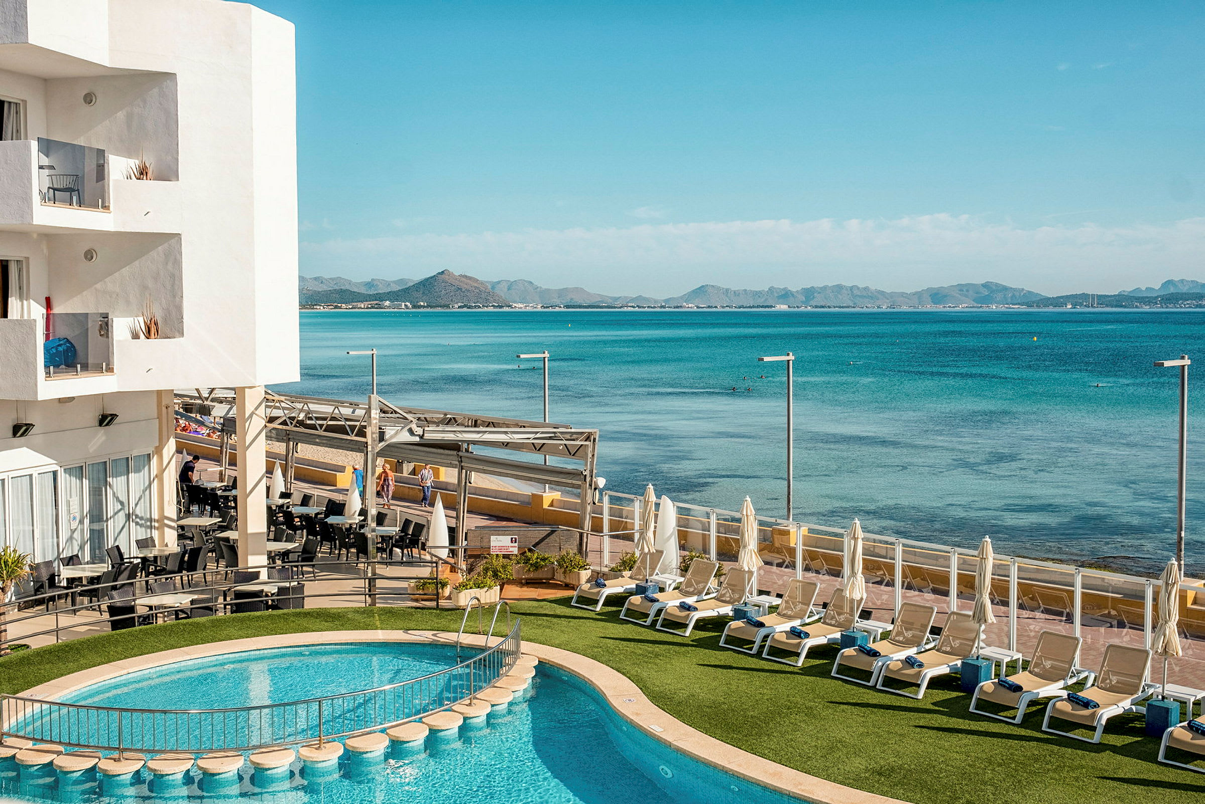 Billedet viser en hotelpool med udsigt over havet og bjergene i baggrunden. (AI-genereret tekst)