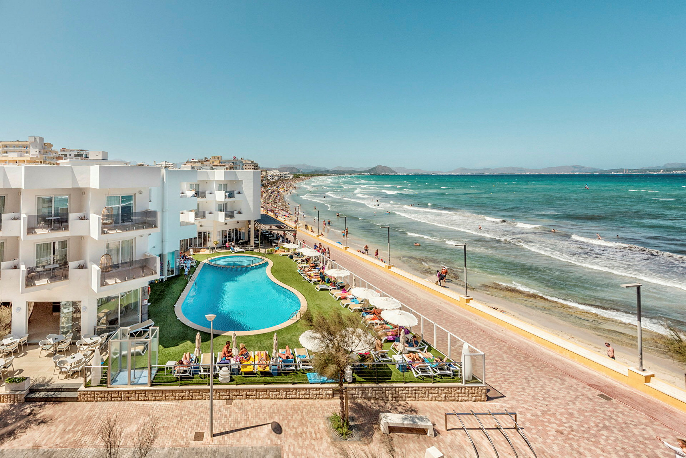 Hotelbygning og poolområde ved strandpromenade og sandstrand. (AI-genereret tekst)