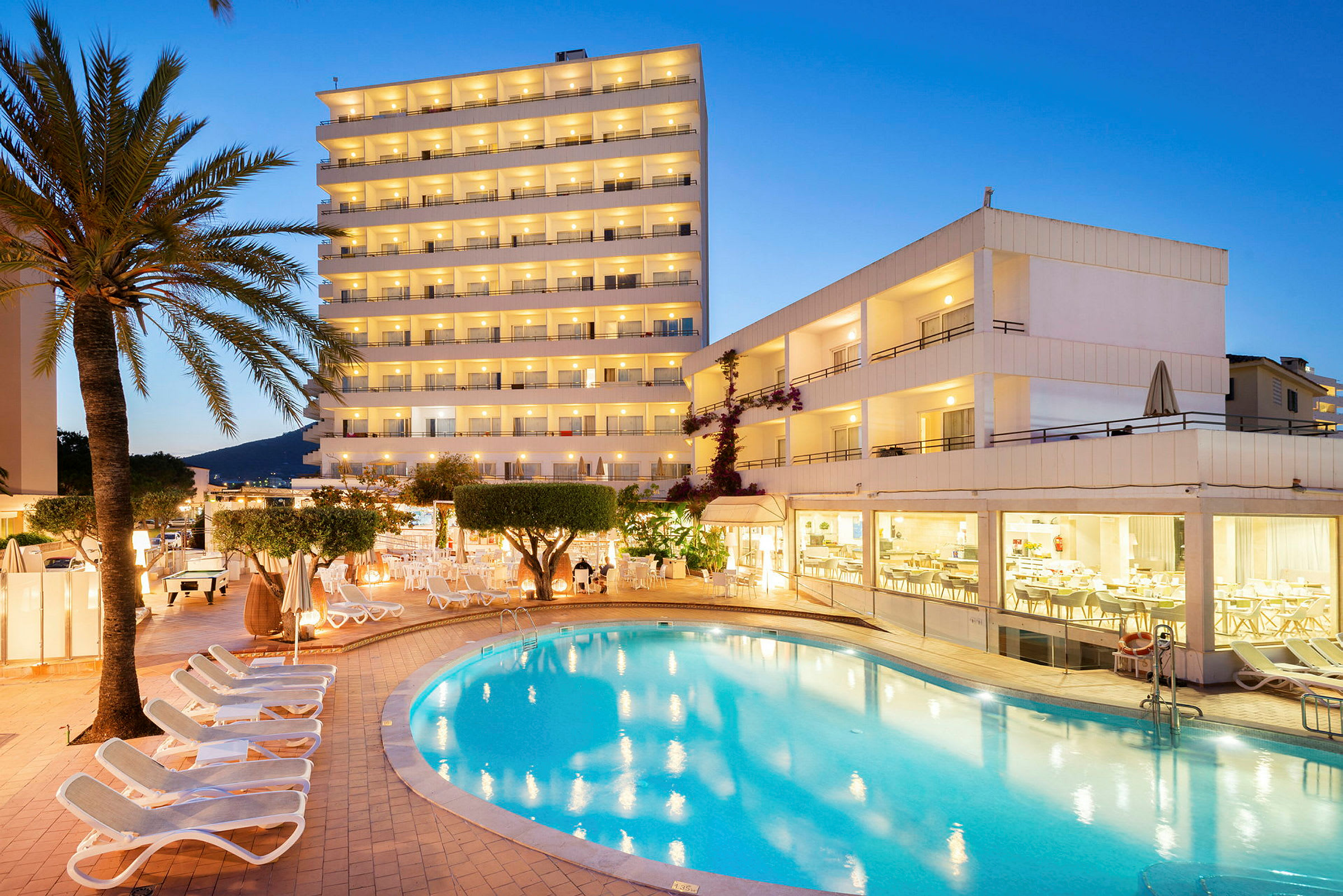 Hotelbygning med oplyste altaner, en swimmingpool og liggestole ved skumringen. (AI-genereret tekst)